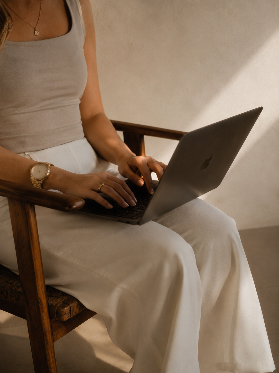 A woman sitting on a wooden chair working on a silver laptop, wearing a beige tank top and white pants, with a gold watch and necklace.