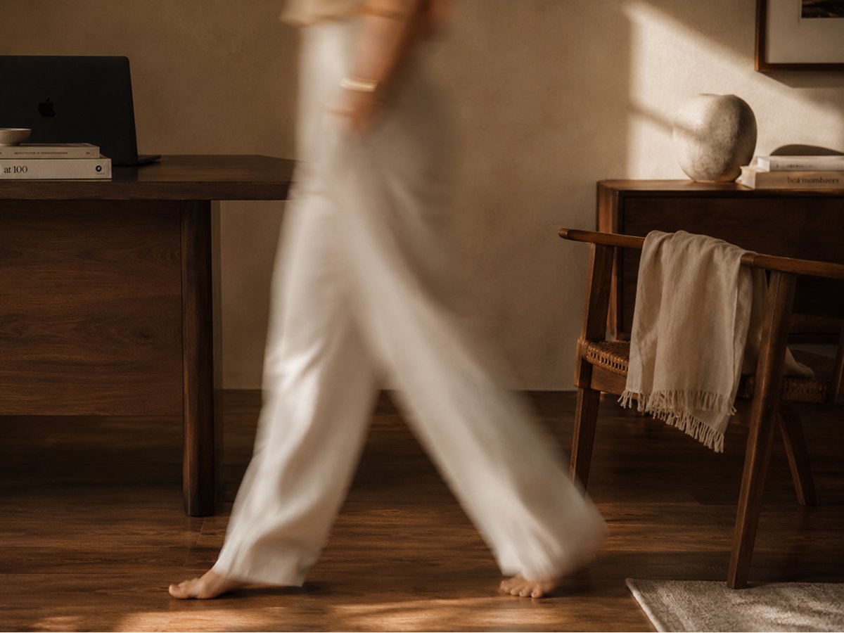 Part of a person walking barefoot in a room with wooden furniture, a desk with books and a laptop, and a wooden chair with a scarf draped over it.