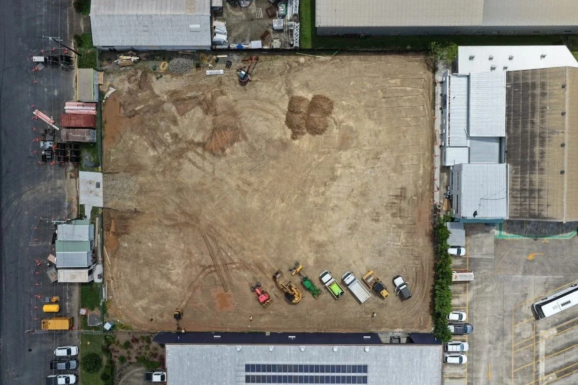 An empty construction site with several small construction vehicles parked along the bottom edge, surrounded by buildings and a parking lot.