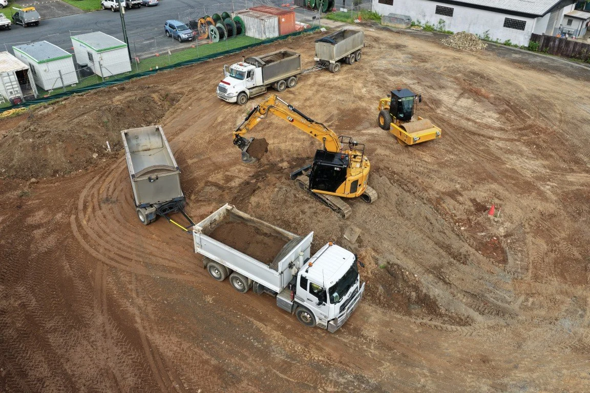 Construction site with a yellow excavator digging soil, three dump trucks, and a yellow compactor on cleared ground.