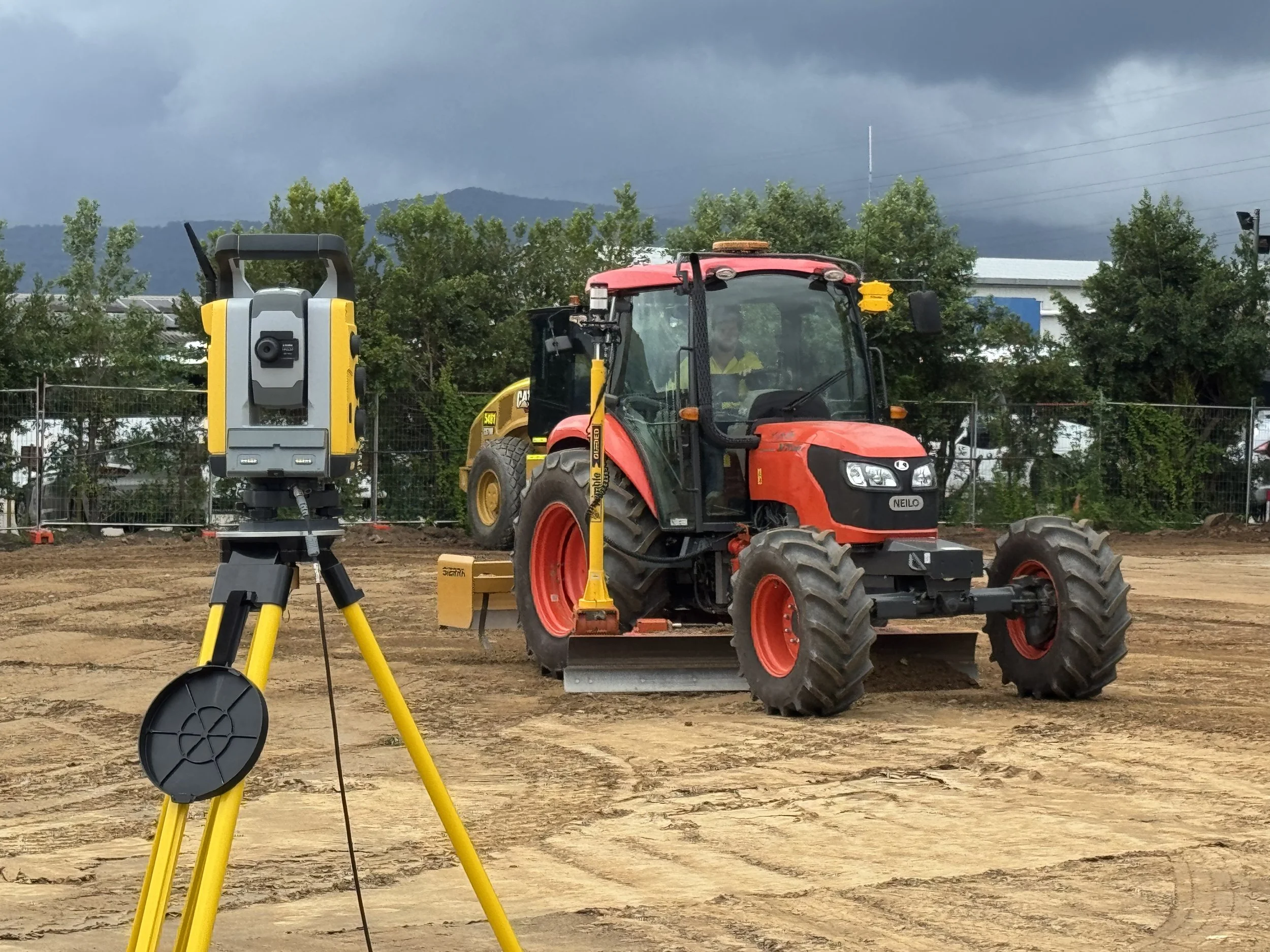 A construction site with a red tractor, a surveying equipment on a tripod, and a cloudy sky in the background.