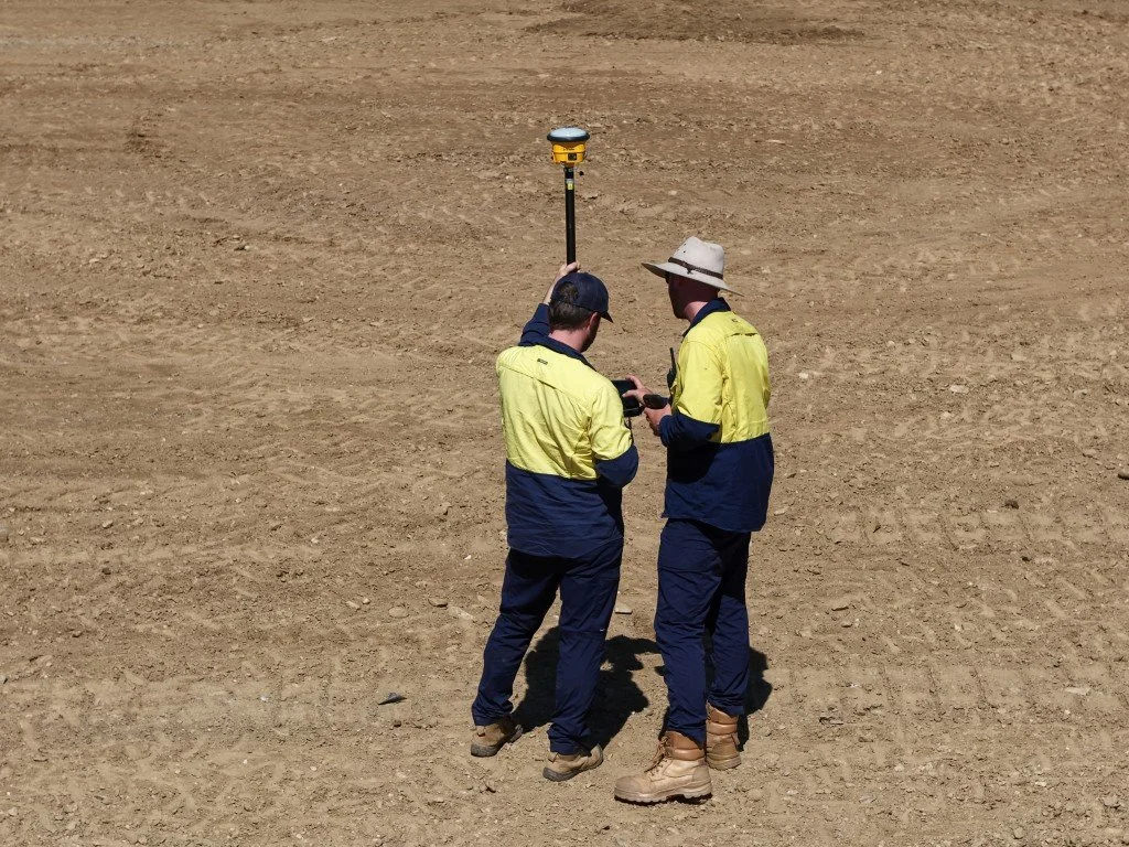 Two workers in high-visibility clothing inspecting equipment on a vast, empty dirt field.