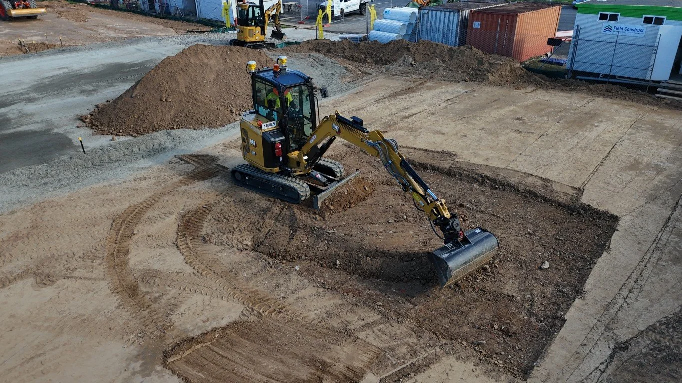 A construction site with a small excavator digging into the dirt. The area includes piles of soil and construction materials, with a backdrop of shipping containers and portable buildings.