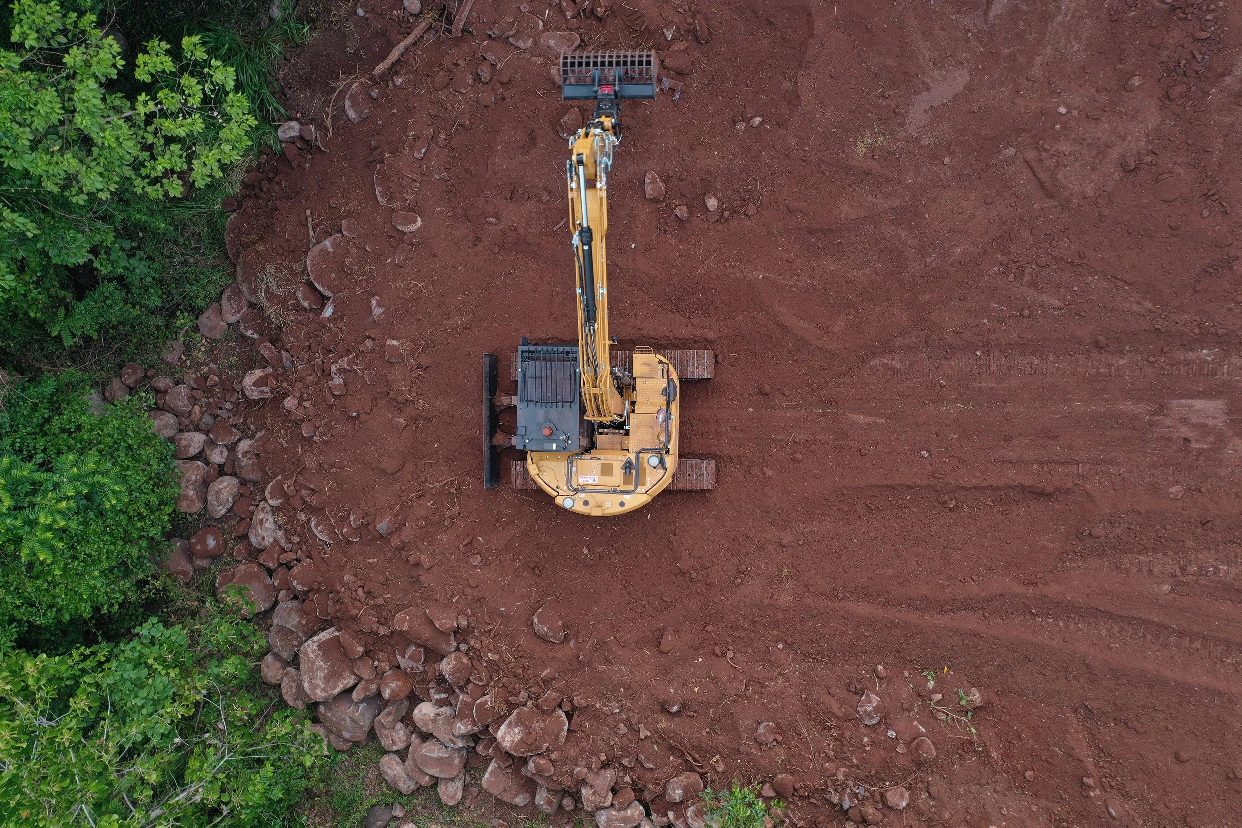 An aerial view of a construction excavator working on a patch of red dirt, with green trees along one side.