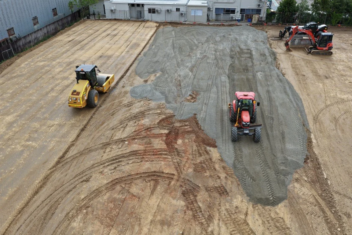 Construction site with machinery preparing the ground for paving, including a yellow roller, a red tractor, and another small excavator, with partially cleared and leveled dirt areas.