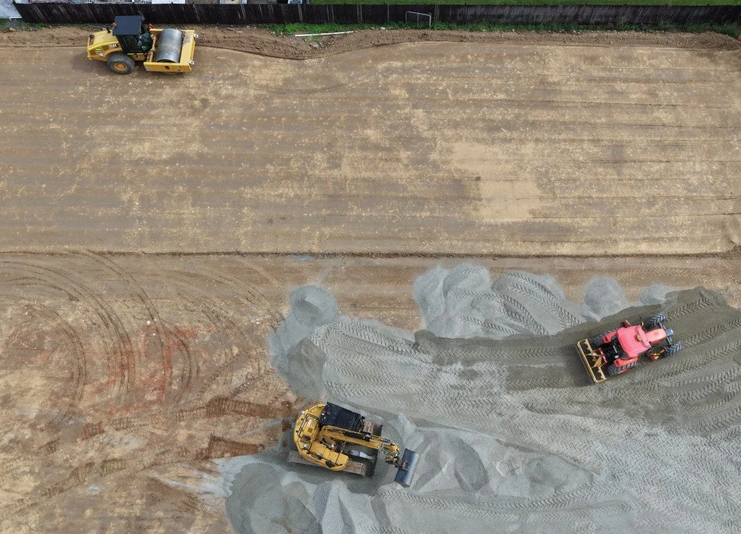 Three construction vehicles, a Cat roller, a red tractro grader and a cat 315 excavator, leveling and compacting dirt on a large construction site.