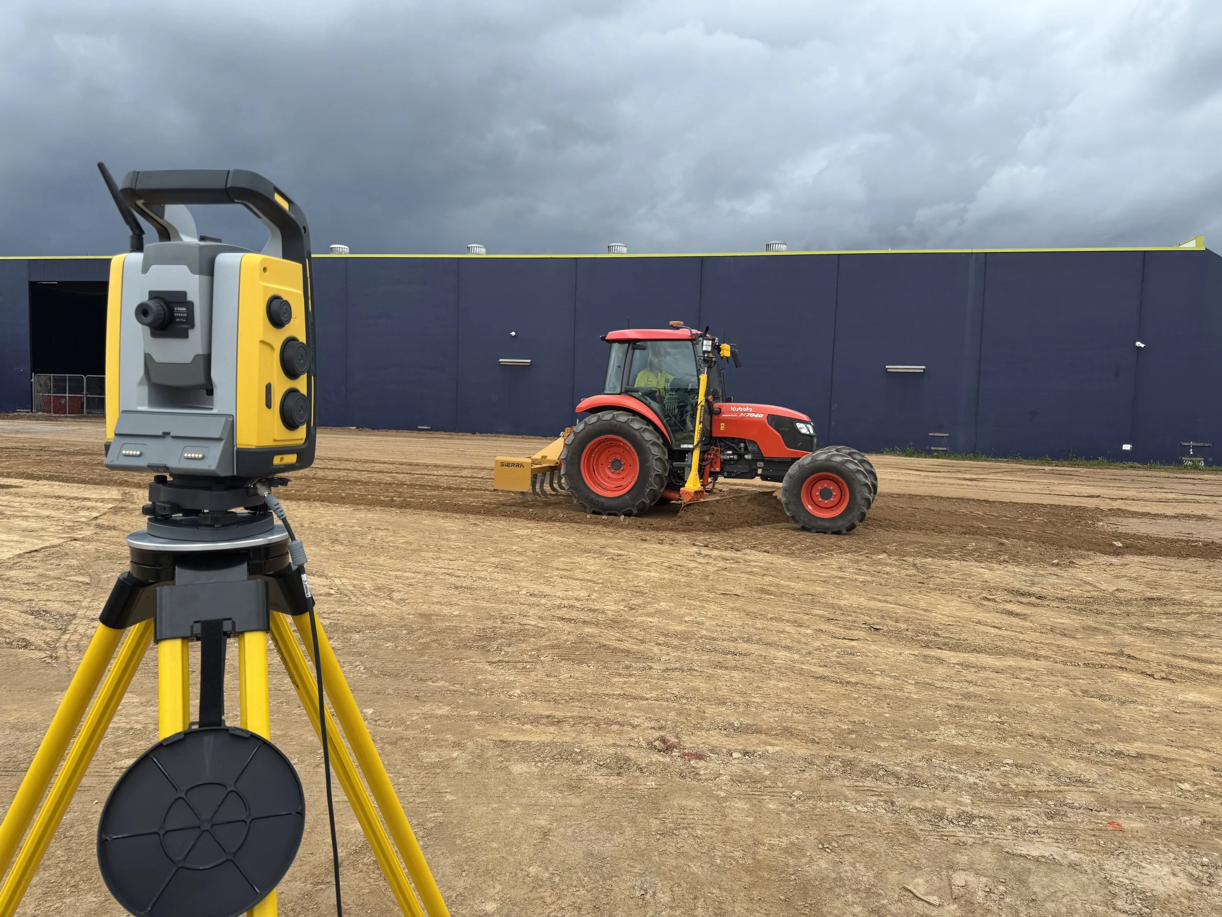 A surveying level mounted on a tripod in the foreground, with a red tractor working on soil in the background, set against a cloudy sky and a dark blue industrial building.