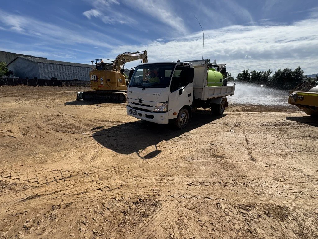 Construction site with a white water tanker truck and a yellow excavator on dirt ground under a partly cloudy sky.