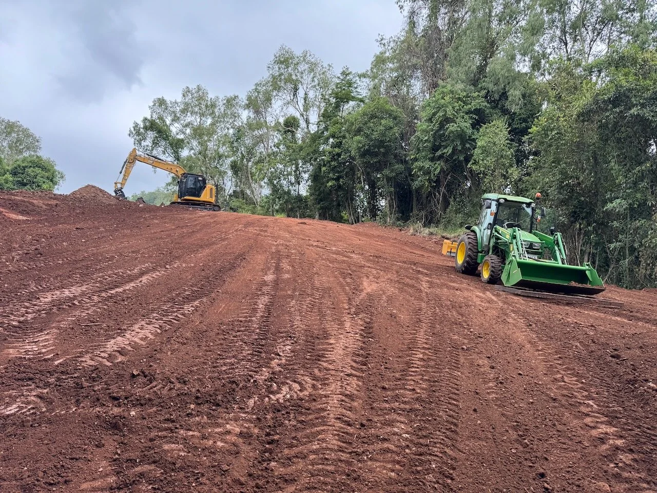 Two construction vehicles working on a dirt slope, with one excavator at the top and a bulldozer at the bottom, amid trees and cloudy sky.