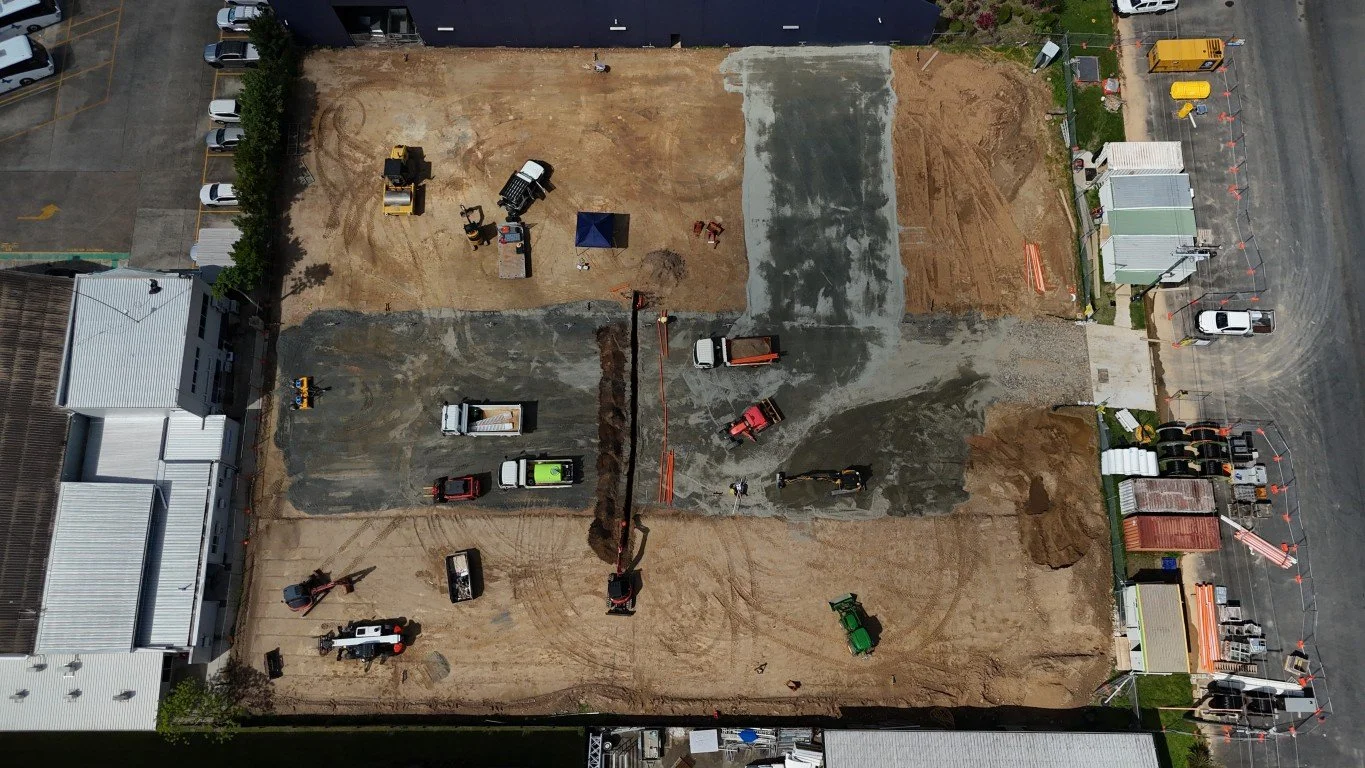 Top-down view of a construction site with machinery, trucks, and construction materials, bordered by buildings and parking areas.