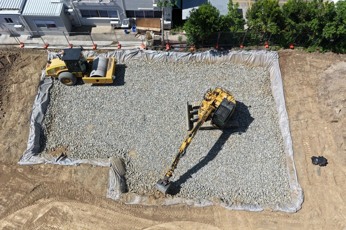 Construction site with a large rectangular area filled with gravel, surrounded by a fabric barrier. There is a yellow compact excavator with a long arm and bucket, and a small yellow road roller nearby. The site is next to a building with a blue exterior and some trees.