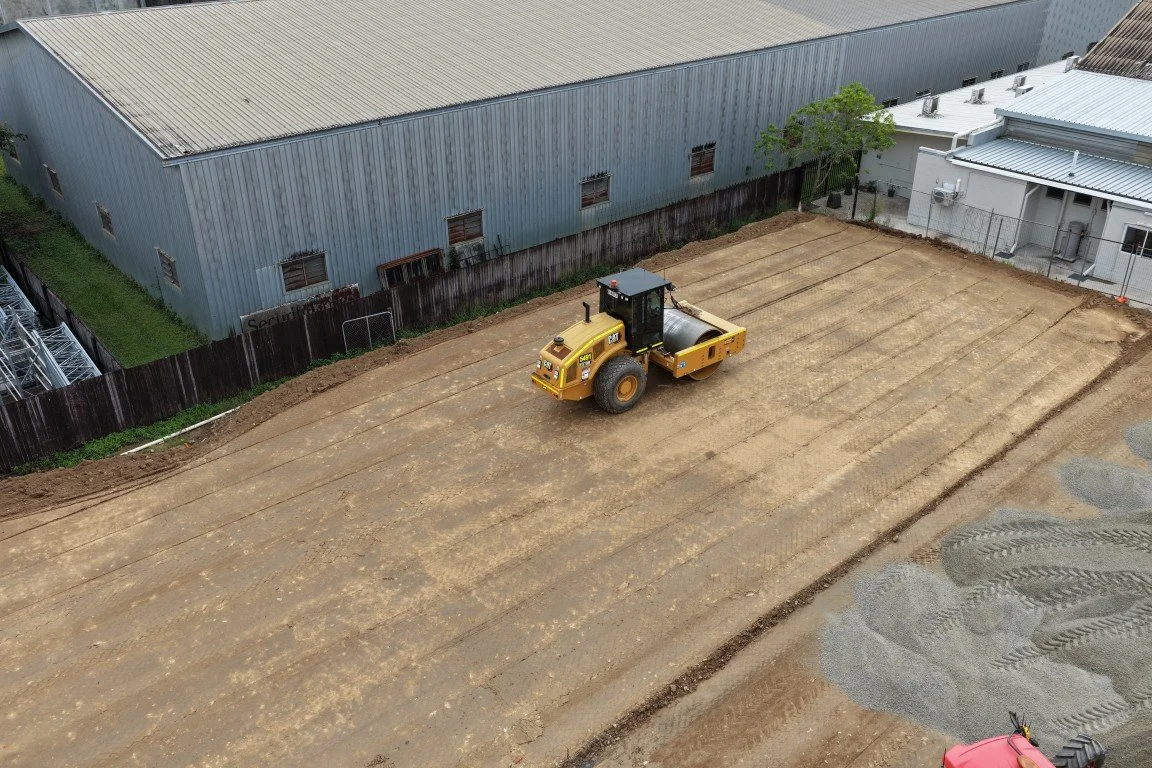 Construction site with a yellow steamroller on leveled dirt ground, adjacent to industrial buildings and a small tree.