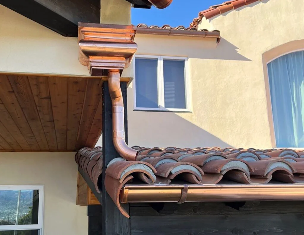 Close-up of a house's roof with red clay tiles, copper gutters, and downspouts, with a cream-colored wall and window with drawn blinds in the background.