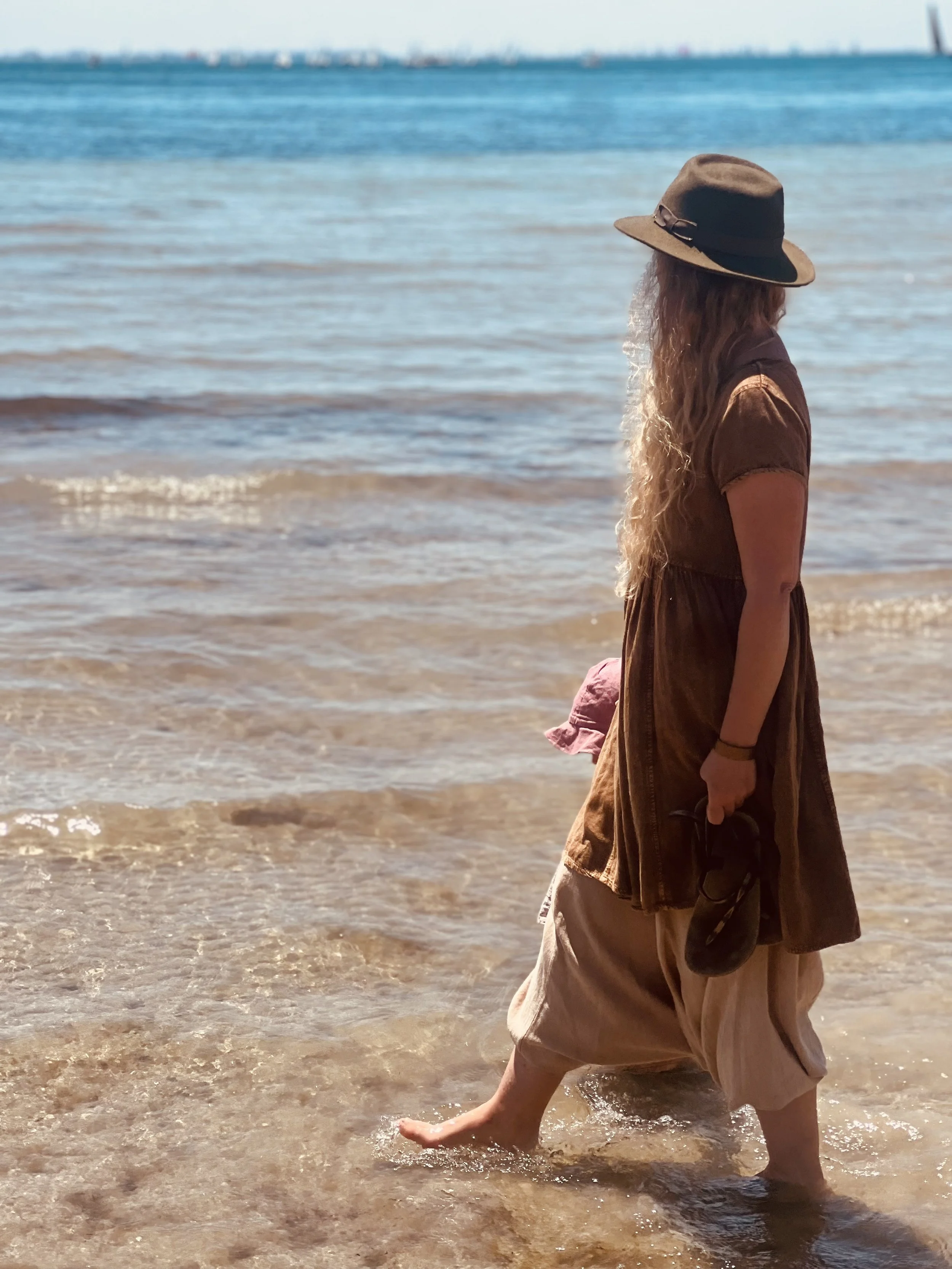 A woman with long curly hair wearing a brown dress and a wide-brimmed hat walking in the shallow water at the beach, holding a pink hat and sunglasses.