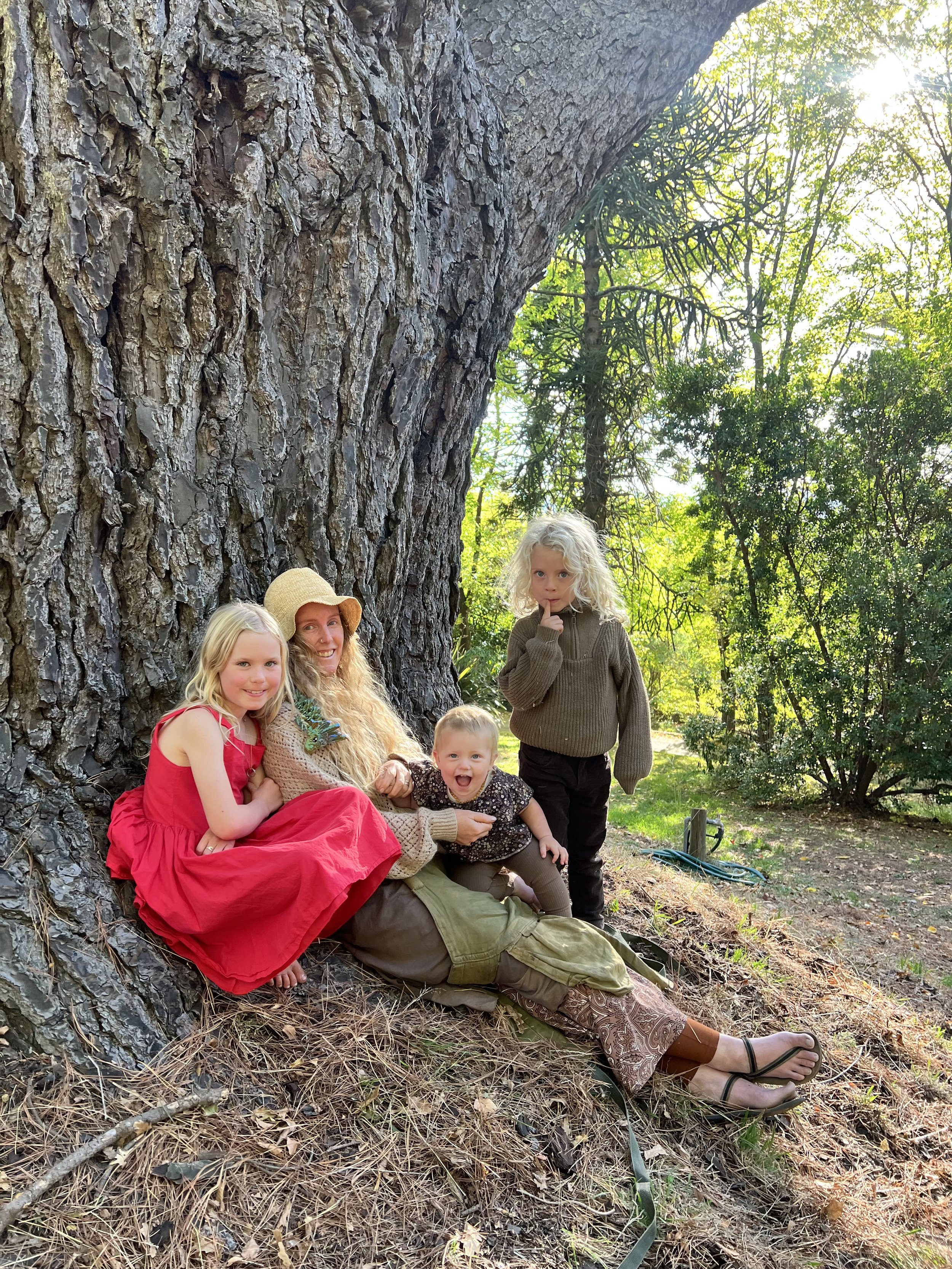 A woman and children sitting under a large tree in a park, with sunlight filtering through the leaves, some children are sitting on her lap and others nearby, all smiling or looking curious.