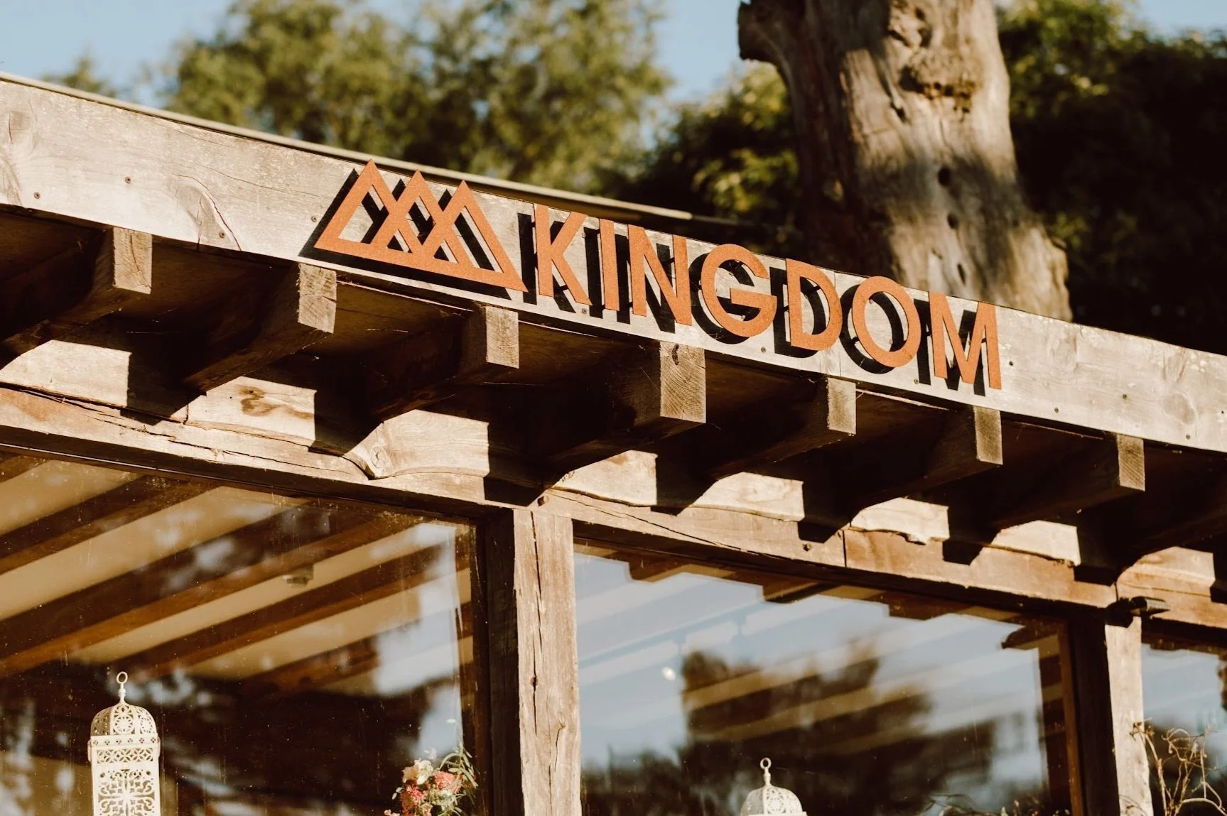 Wooden structure with a sign that reads 'KINGDOM' in orange letters, with a triangular mountain design to the left of the text. The background includes a tree and blue sky, and the interior shows hanging decorative lanterns.