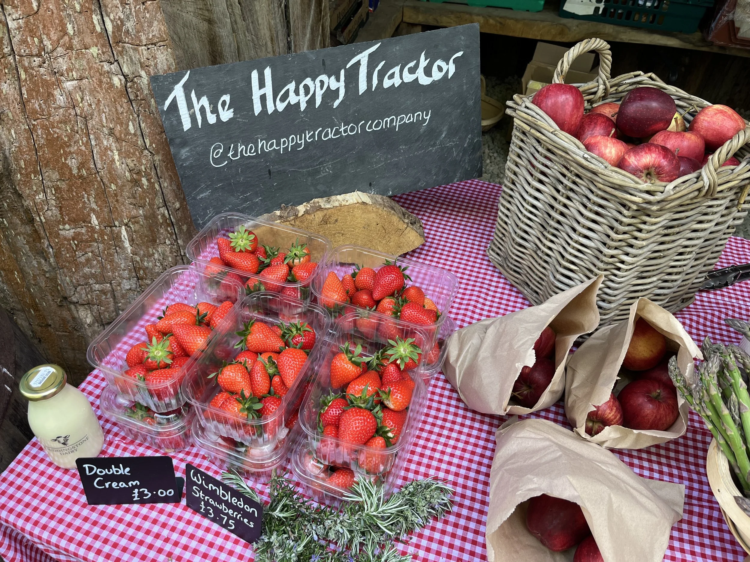 Fresh strawberries in clear plastic containers, red apples in a wicker basket, a small bottle of milk, and handwritten price signs on a red and white checkered tablecloth at a farmers market stand.