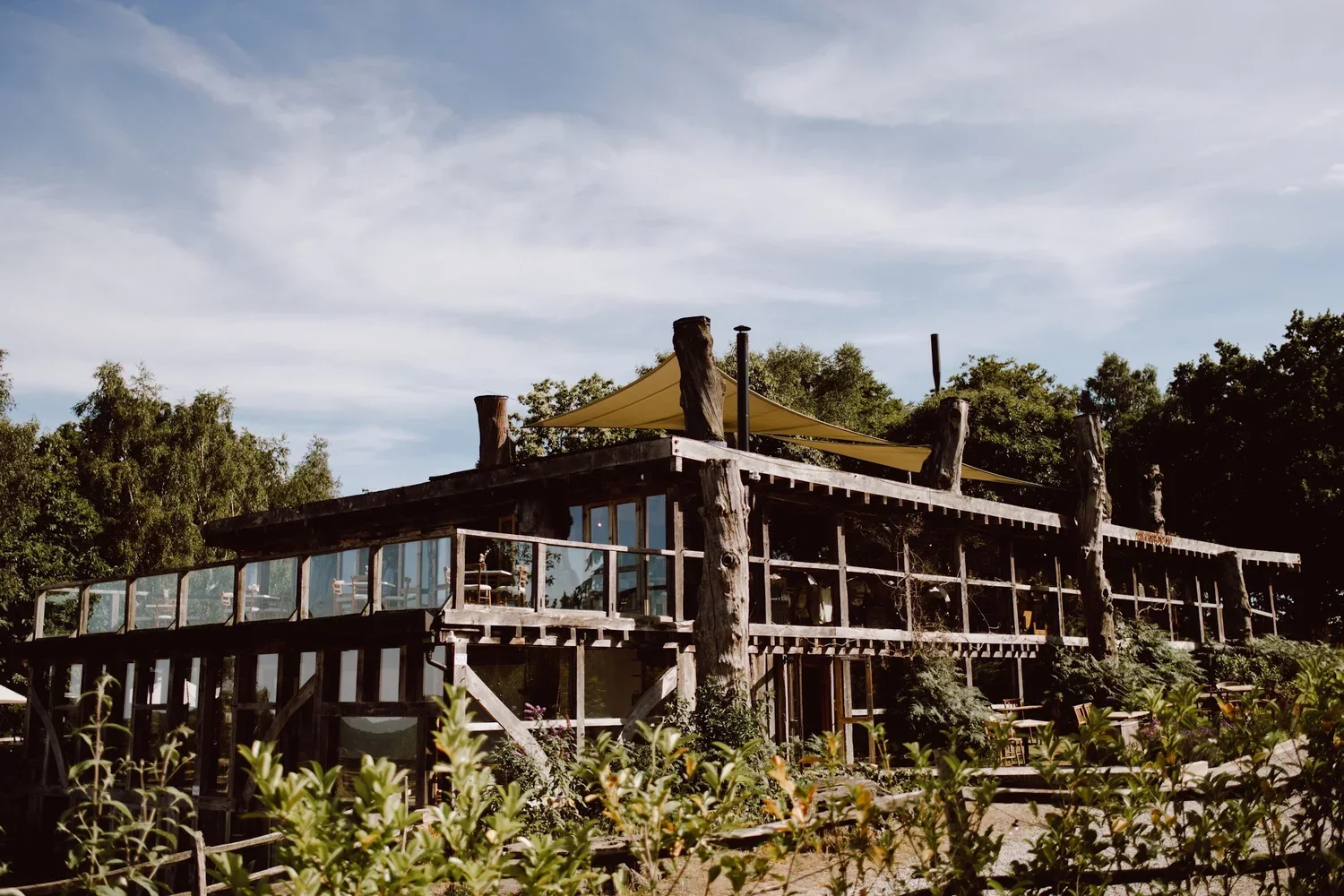 A rustic, multi-level wooden building with large glass windows, situated among trees and greenery, featuring an outdoor terrace with a sunshade on the top level, under a partly cloudy sky.