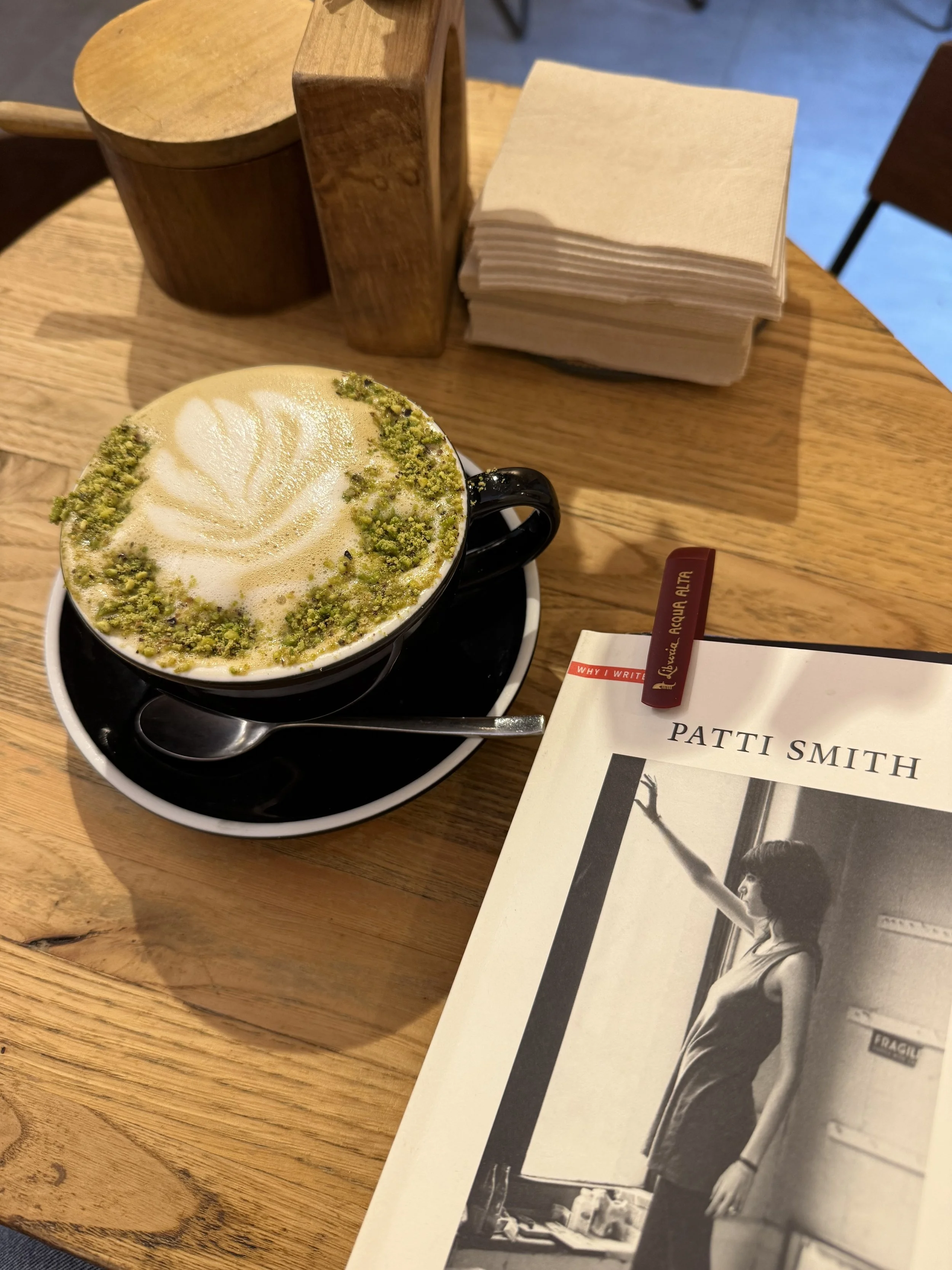 A cup of coffee with foam art and sprinkled pistachios on top, placed on a wooden table next to a book and a napkin holder.