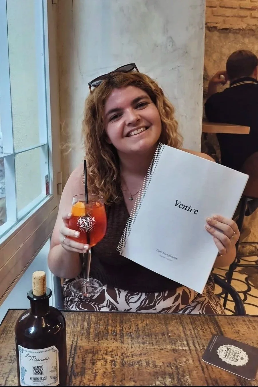 Woman with curly hair smiling, holding a book labeled 'Venice', and a glass of Aperol Spritz with an orange slice, seated at a rustic wooden table in a restaurant.