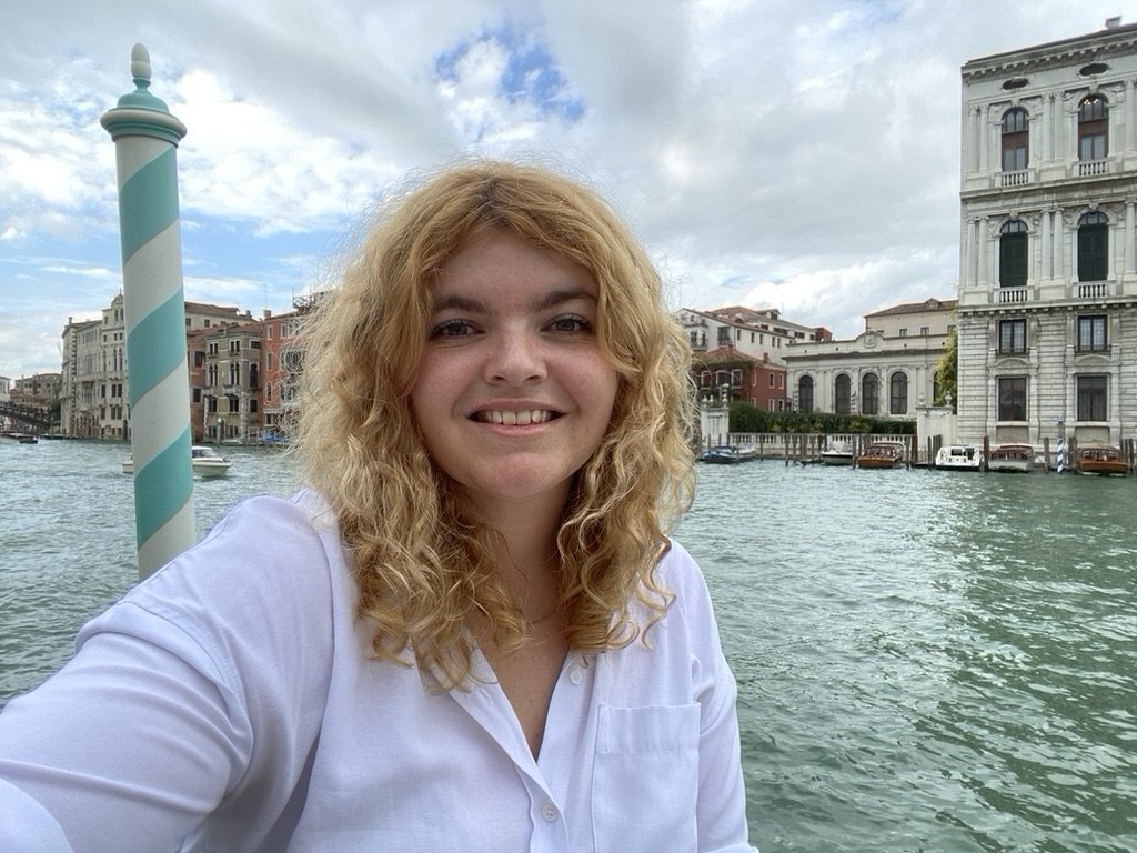 Smiling woman with curly blonde hair taking a selfie on a waterway in Venice, Italy, with historic buildings and boats in the background.