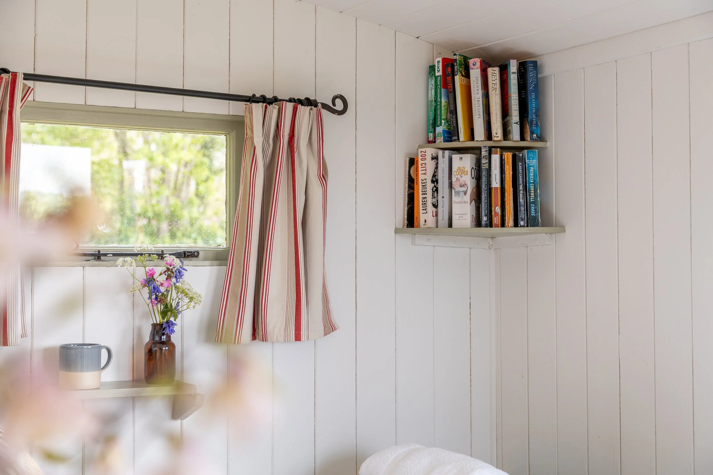 A cozy white-walled room with a small shelf holding a vase of colorful flowers, a white mug, and a brown glass bottle. A window with red and white striped curtains allows natural light to brighten the space. Two white bookshelves with a variety of bo