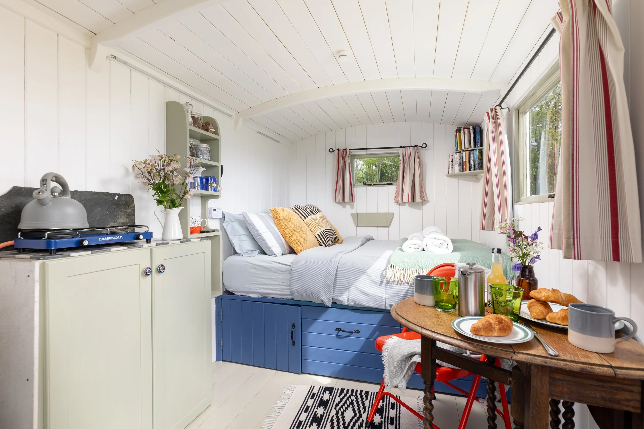 A cozy bedroom with white paneled walls and ceiling, a bed with striped pillows, small window with striped curtains, a green shelf with glassware and jars, a small table with breakfast items including croissants, mugs, and a plant, and a black-and-wh