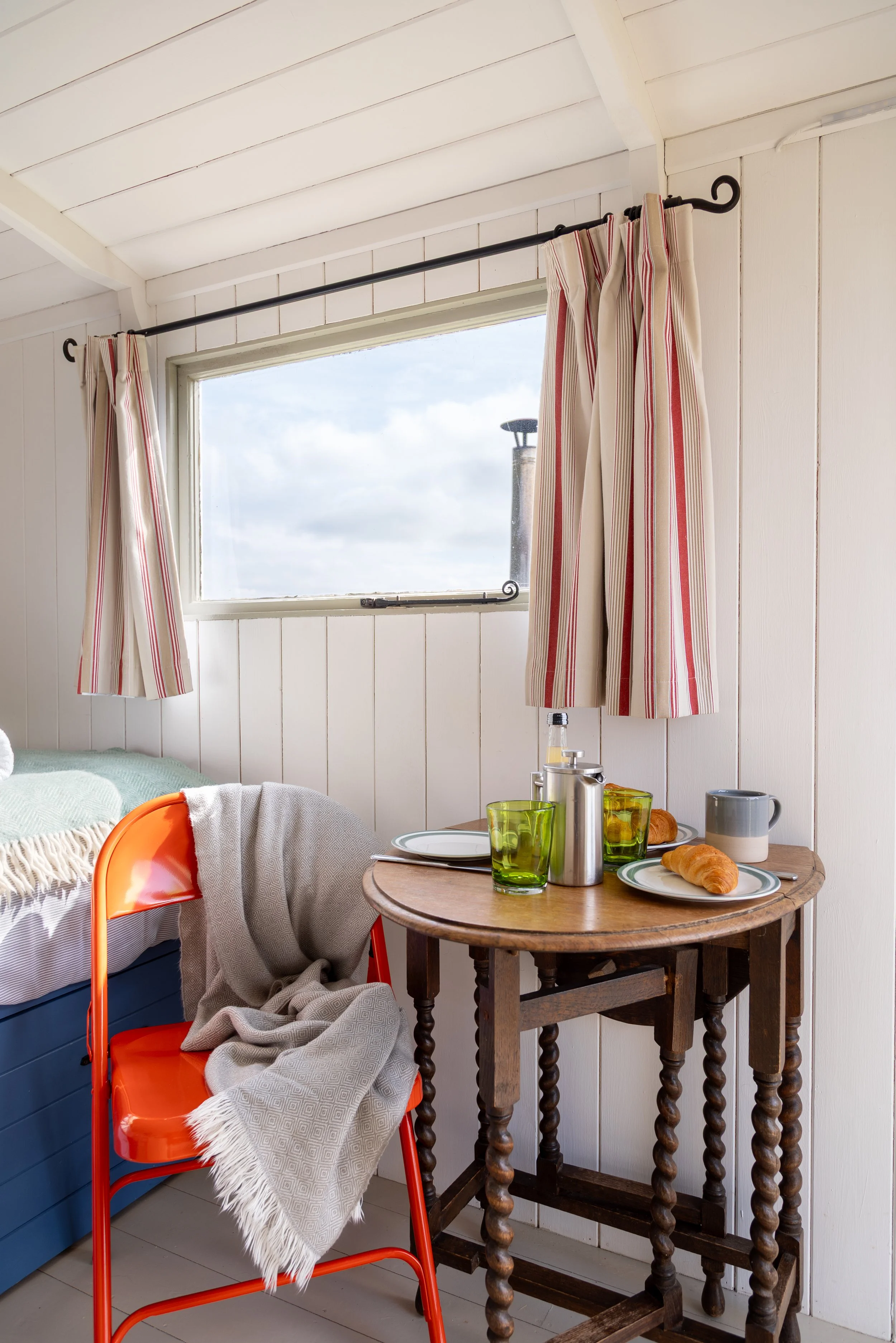 A cozy indoor scene with a window on white wooden walls, red and white striped curtains, a wooden table set with breakfast items including croissants, green glasses, a silver thermos, and plates, alongside a red chair with a beige blanket draped over