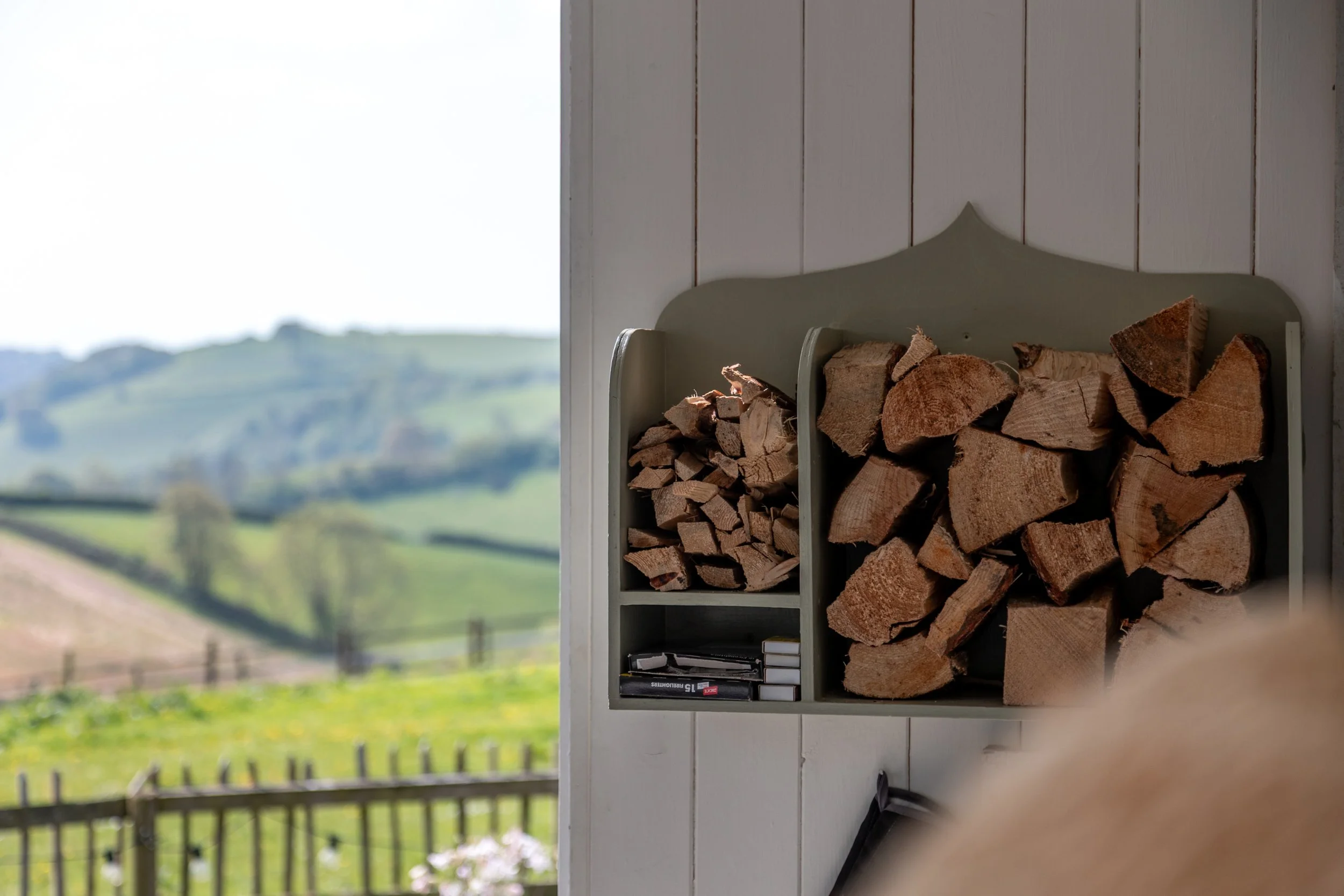 A wood storage rack filled with chopped firewood, positioned outdoors near a white wooden wall with a scenic view of green rolling hills and farmland in the background.