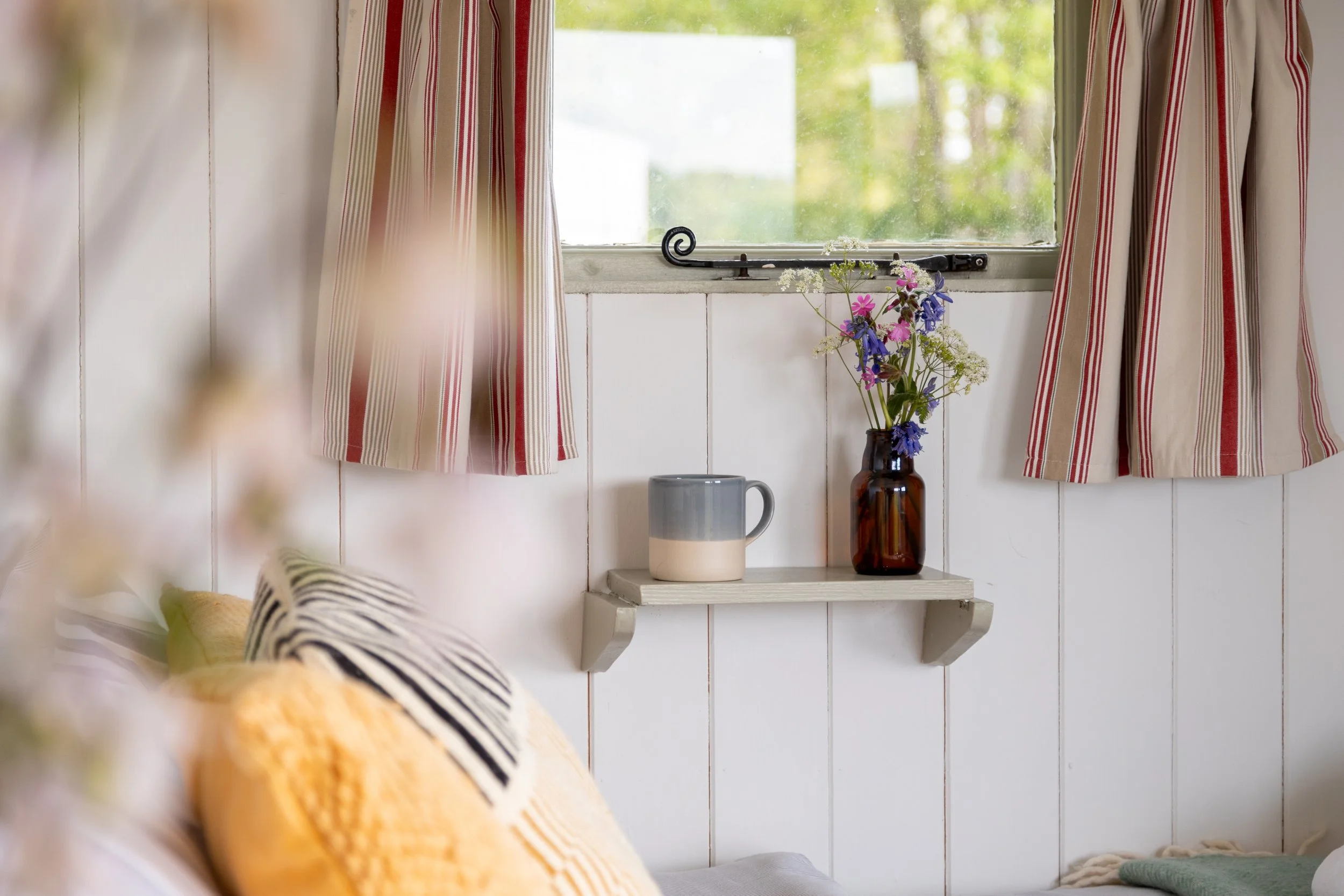 A window with cream-colored curtains with red and beige stripes, a small white shelf with a brown vase holding a bouquet of pink, purple, and white flowers, and a tall ceramic mug with a gradient beige and gray design, in a cozy room with white panel