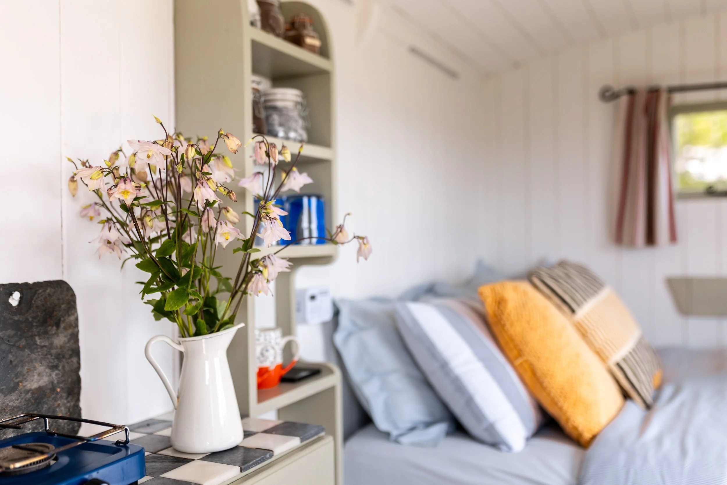 A white pitcher holding pink and white flowers placed on a kitchen counter. In the background, there is a shelf with kitchen items and a bed with pillows in a small room with a window.