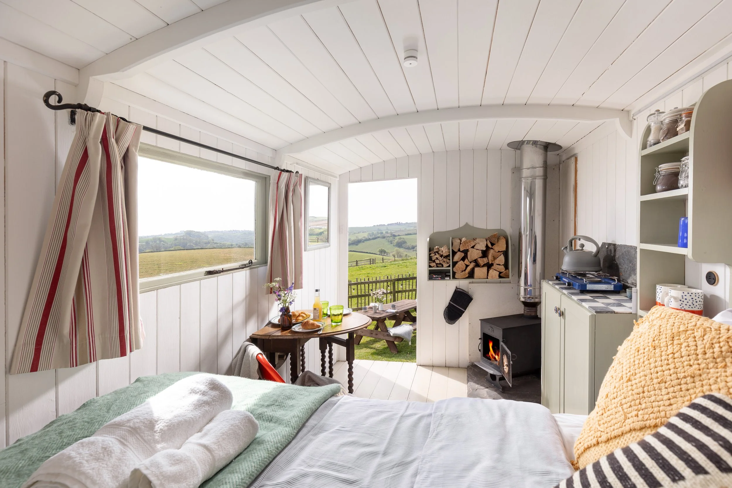 Cozy interior of a sunlit room with white wooden walls and ceiling, large windows showing a green countryside landscape, a small dining table with chairs and breakfast items, a wood-burning stove with logs, a white shelf with jars and dishes, and a b
