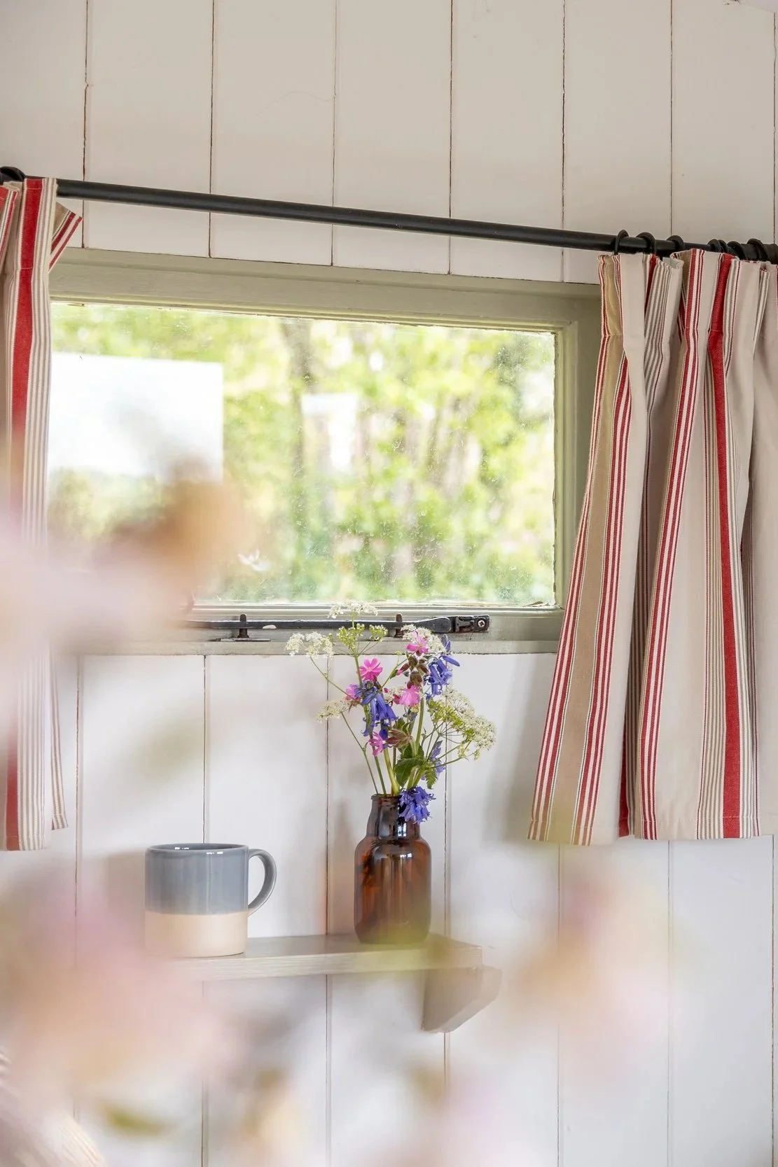 A small window with red and white striped curtains, a brown vase with wildflowers, and a ceramic mug on a shelf inside a white-tiled room.