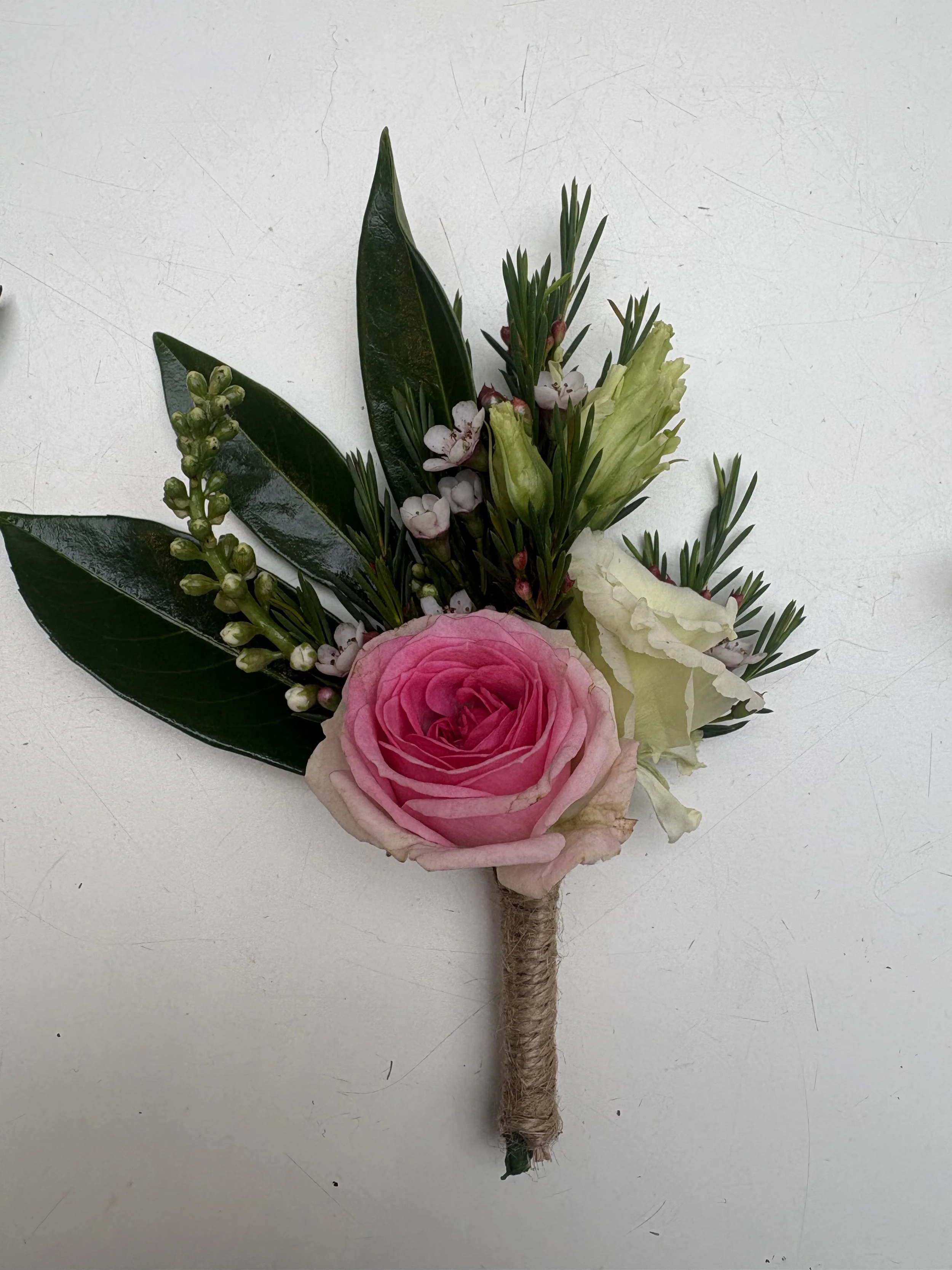 A beautiful button hole for wedding grooms, with a pink rose, white flowers, green foliage, wrapped with string, perfect for a countryside wedding.