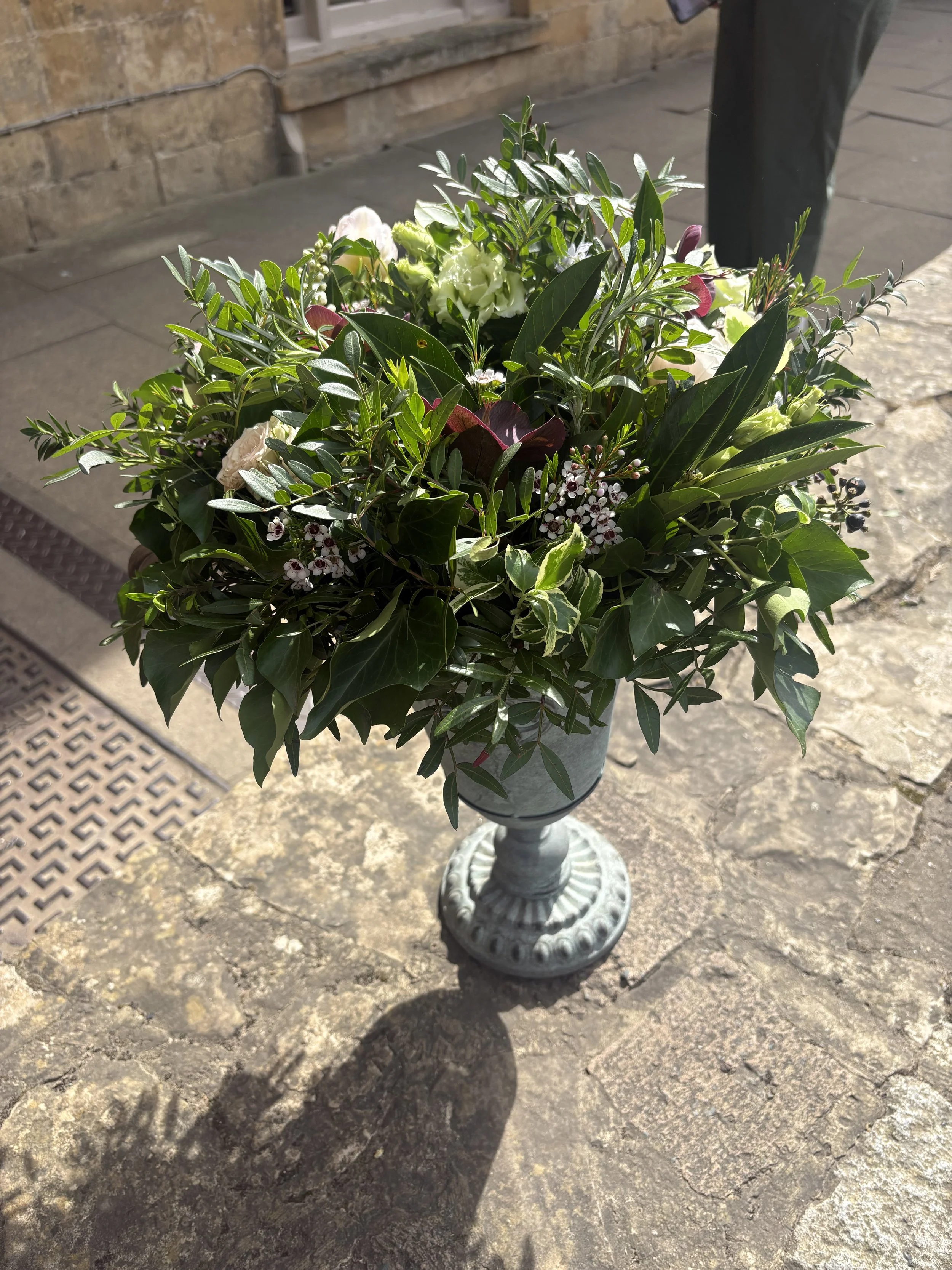 A decorative pedestal urn with a large arrangement of green and white flowers and seasonal foliage.