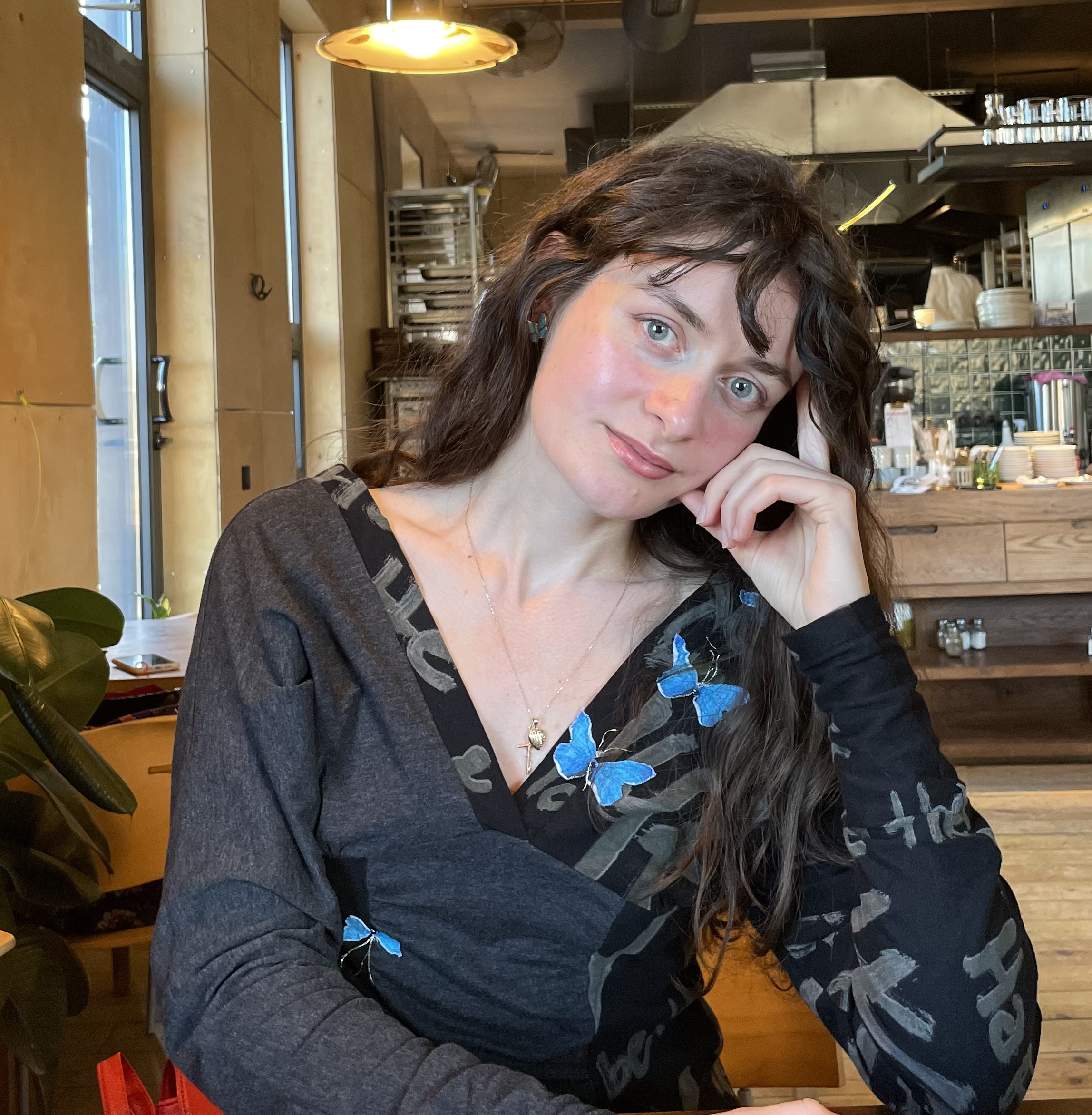 A woman with long, wavy dark hair and blue eyes sitting at a wooden table in a cozy restaurant or cafe, with wooden-paneled walls, large windows, and a kitchen in the background.