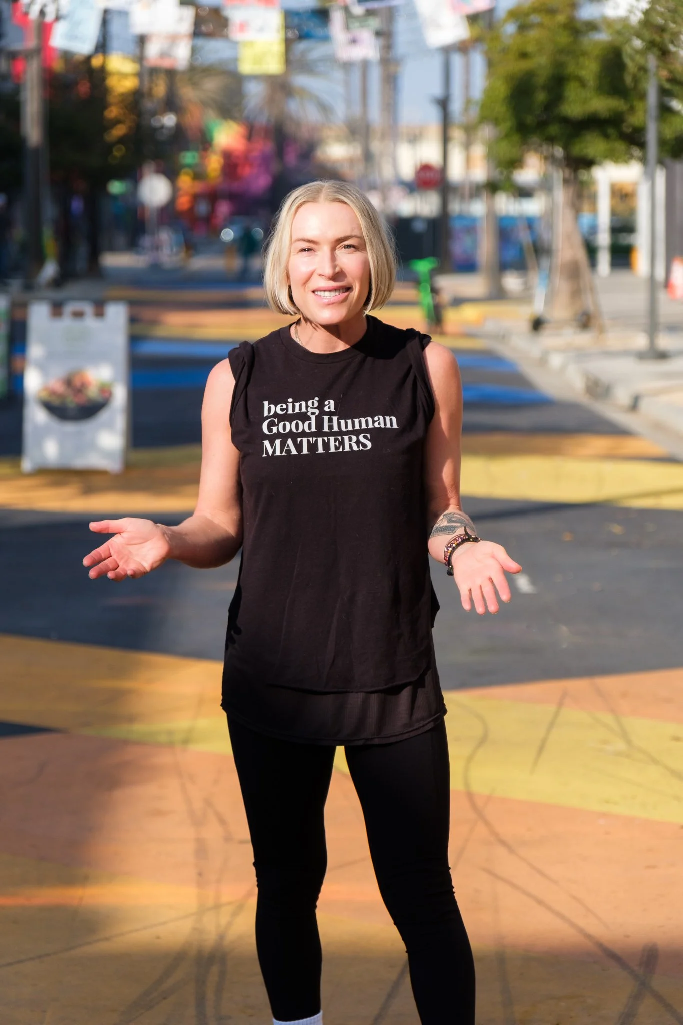 A woman with shoulder-length blonde hair smiling and shrugging her shoulders, standing on a basketball court outdoors on a sunny day, wearing a black sleeveless shirt with white text, black leggings, and bracelets.