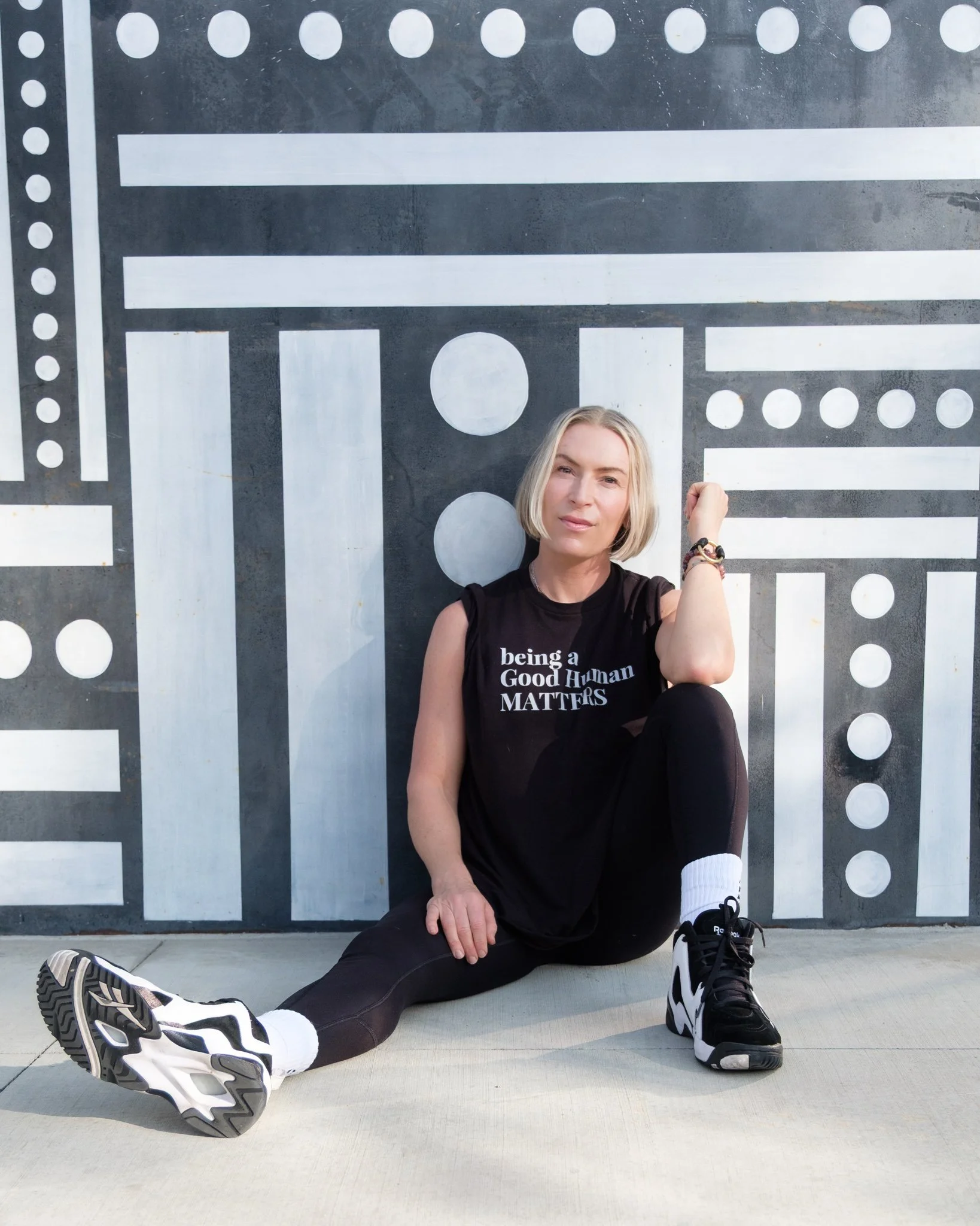 A woman with blonde hair sitting on the ground in front of a black and white geometric mural, wearing a black sleeveless shirt that reads 'being a Good Human MATTERS,' black leggings, white socks, and black and white athletic shoes, with one arm raised in a fist.