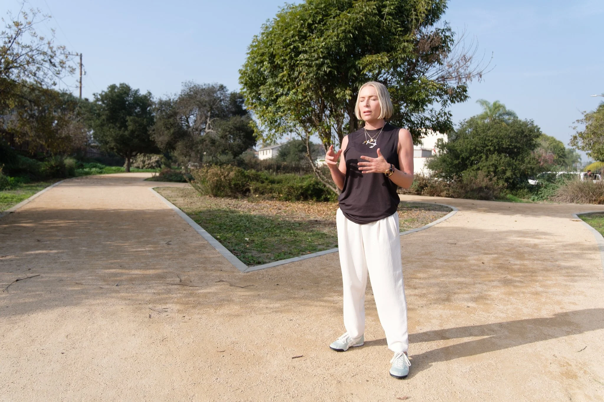 A woman with blonde hair, dressed in a black sleeveless top and white pants, standing on a park pathway with trees and bushes in the background, outdoors on a sunny day.