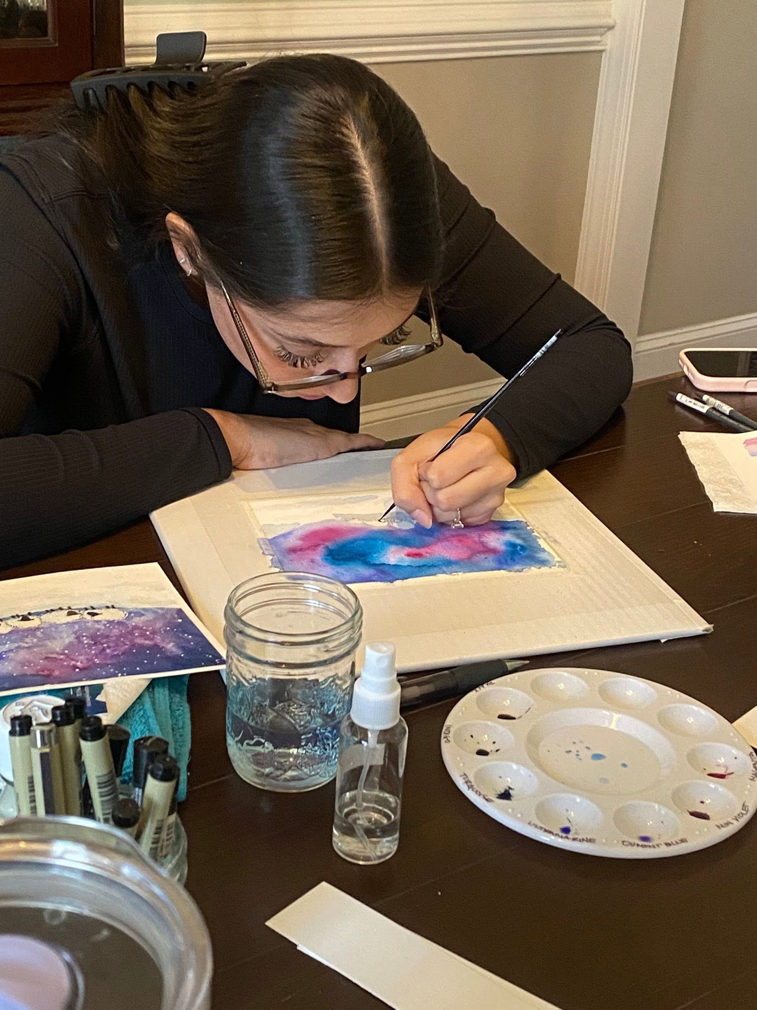 Person with long dark hair and glasses painting watercolor on paper at a table with art supplies and palette.