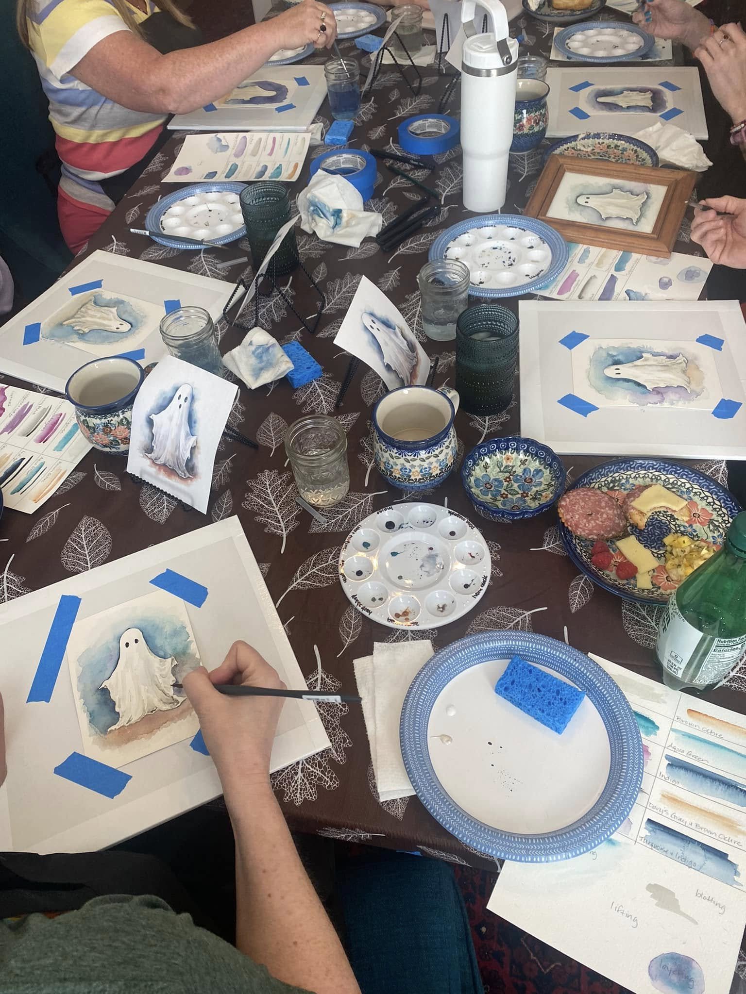 People sitting around a table painting watercolor ghost illustrations on paper. Supplies like paint palettes, brushes, water, painter's tape, and finished ghost artwork are visible.