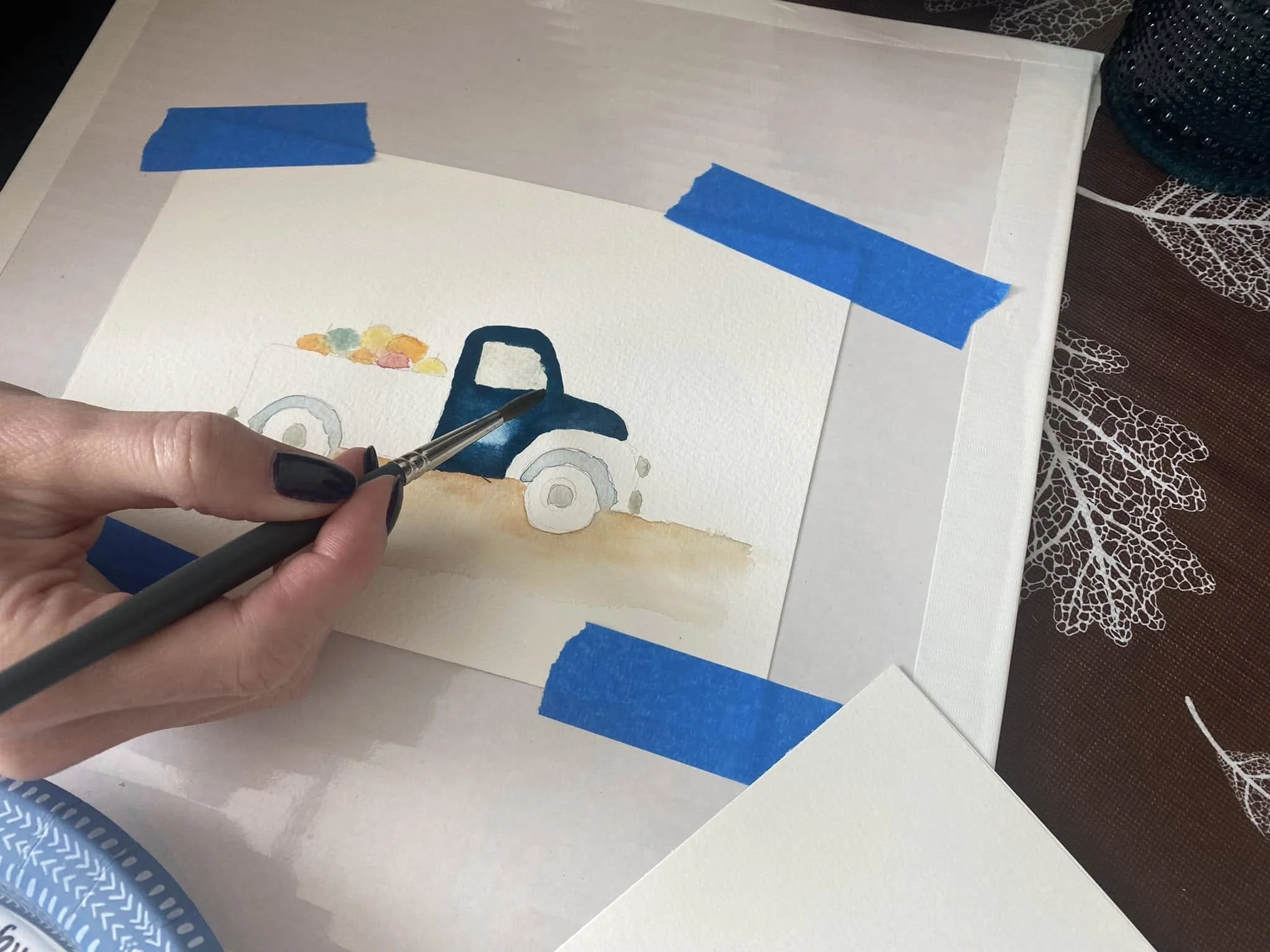 A person holding a paintbrush working on a watercolor painting of a truck with balloons beside it, on a piece of paper taped to a surface with blue painter's tape. There is a lace tablecloth underneath the paper.