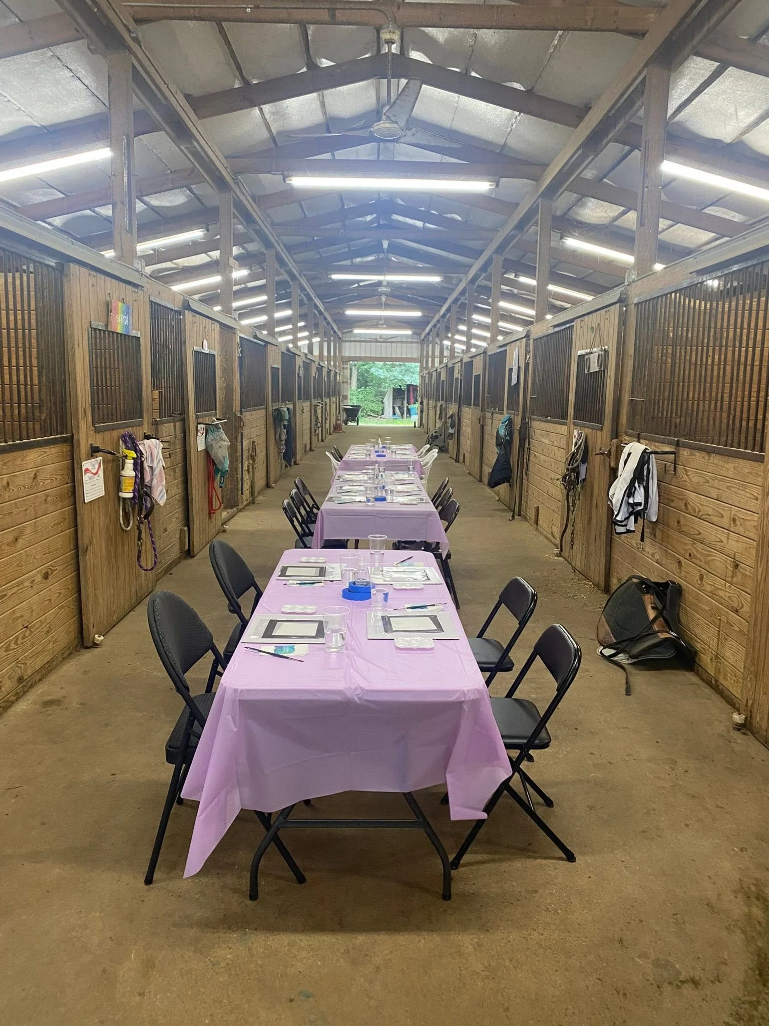 A long dining table covered with a pink tablecloth, set with clear cups and silverware, in an indoor barn with wooden stalls on either side, decorated for a celebration.