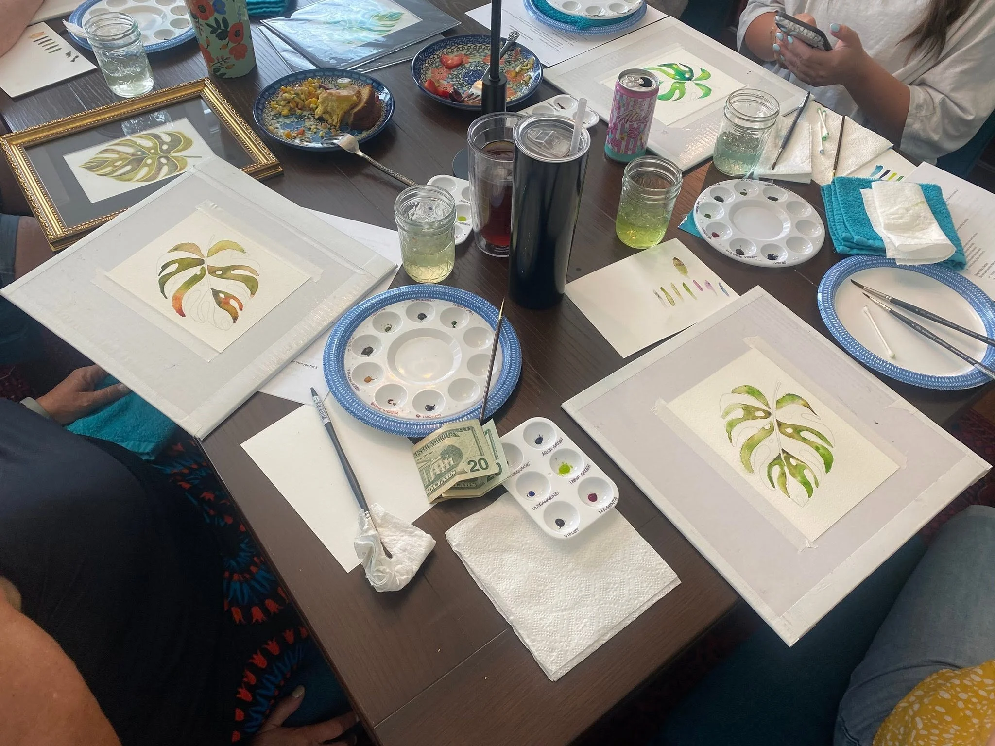 A table with art supplies including watercolor palettes, brushes, paper with painted leaves, glasses of water, and partially eaten slices of cake. People are working on watercolor paintings, and some are using their phones.