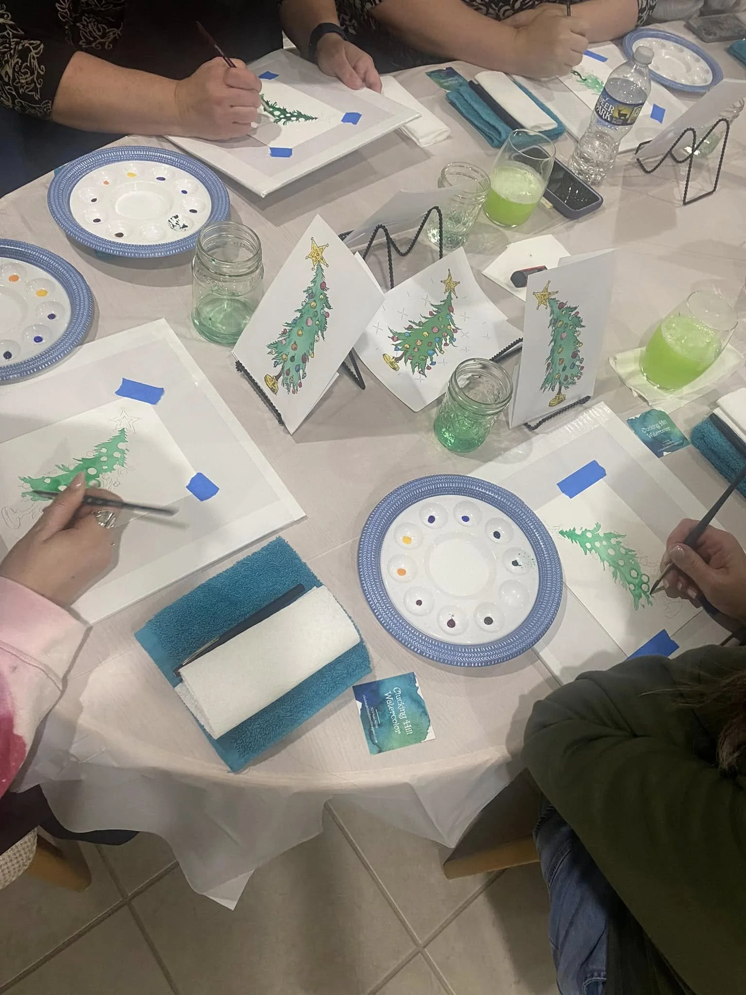 People sitting at a table painting Christmas tree and holiday cards, with water bottles, glasses, paper plates, and art supplies on the table.