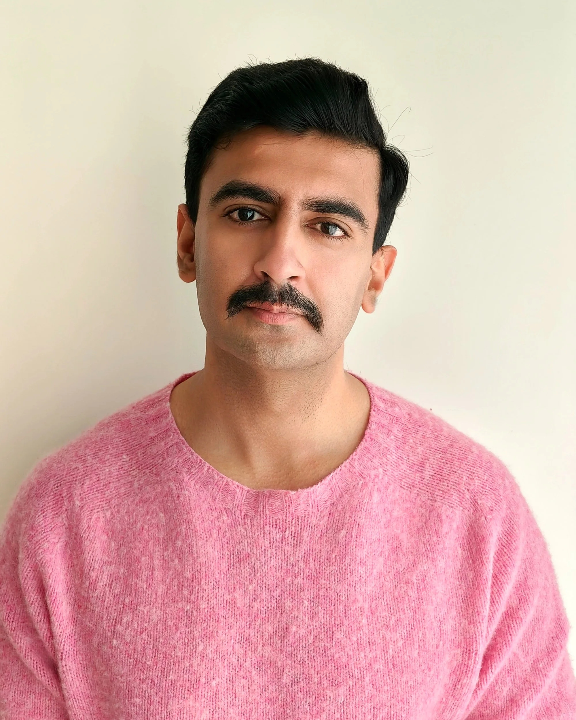 Close-up of a young man with dark hair, mustache, and goatee, wearing a pink sweater, standing against a plain off-white wall.
