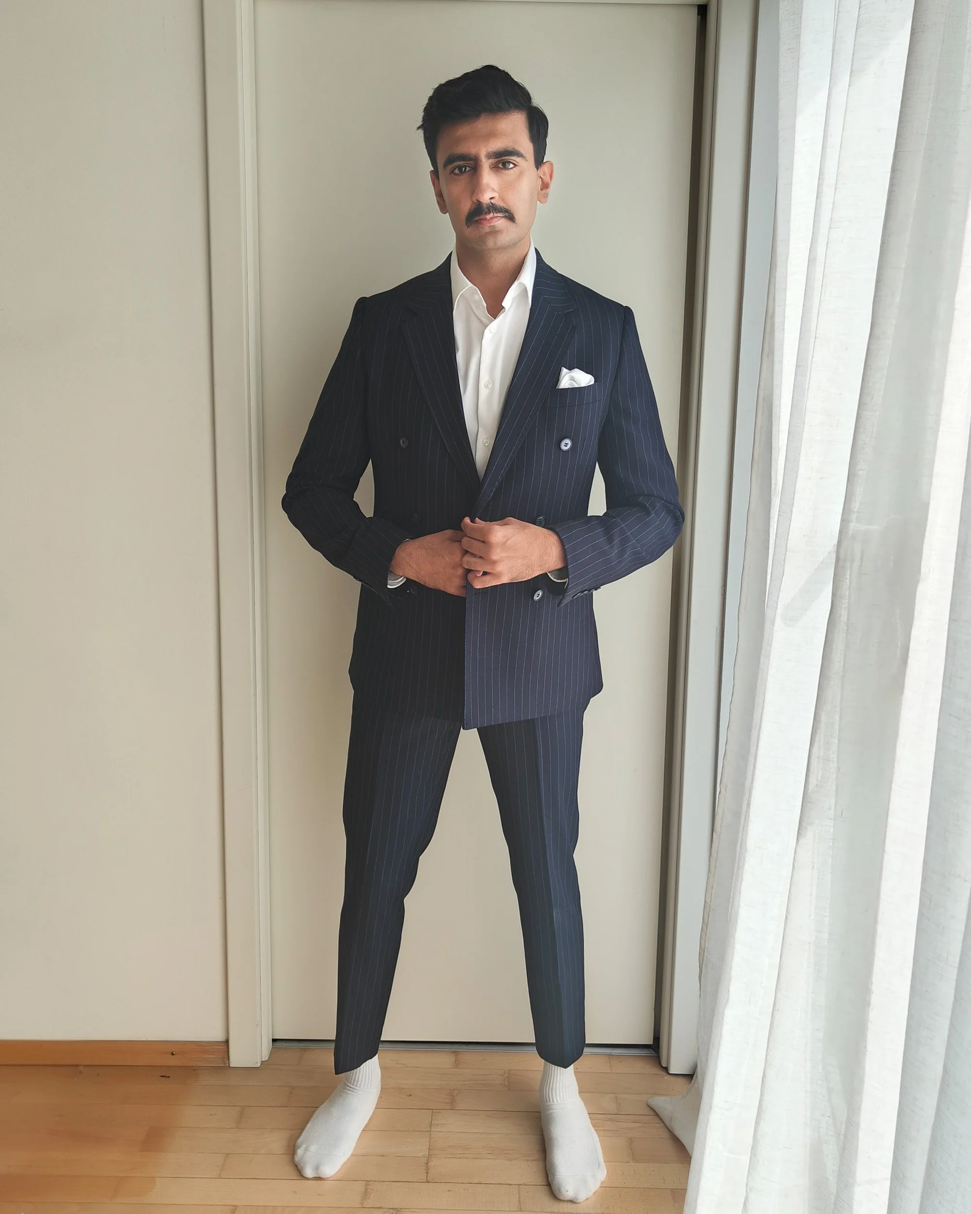 A man in a dark pinstripe suit with a white shirt and pocket square standing inside by a window with curtains, wearing socks instead of shoes.