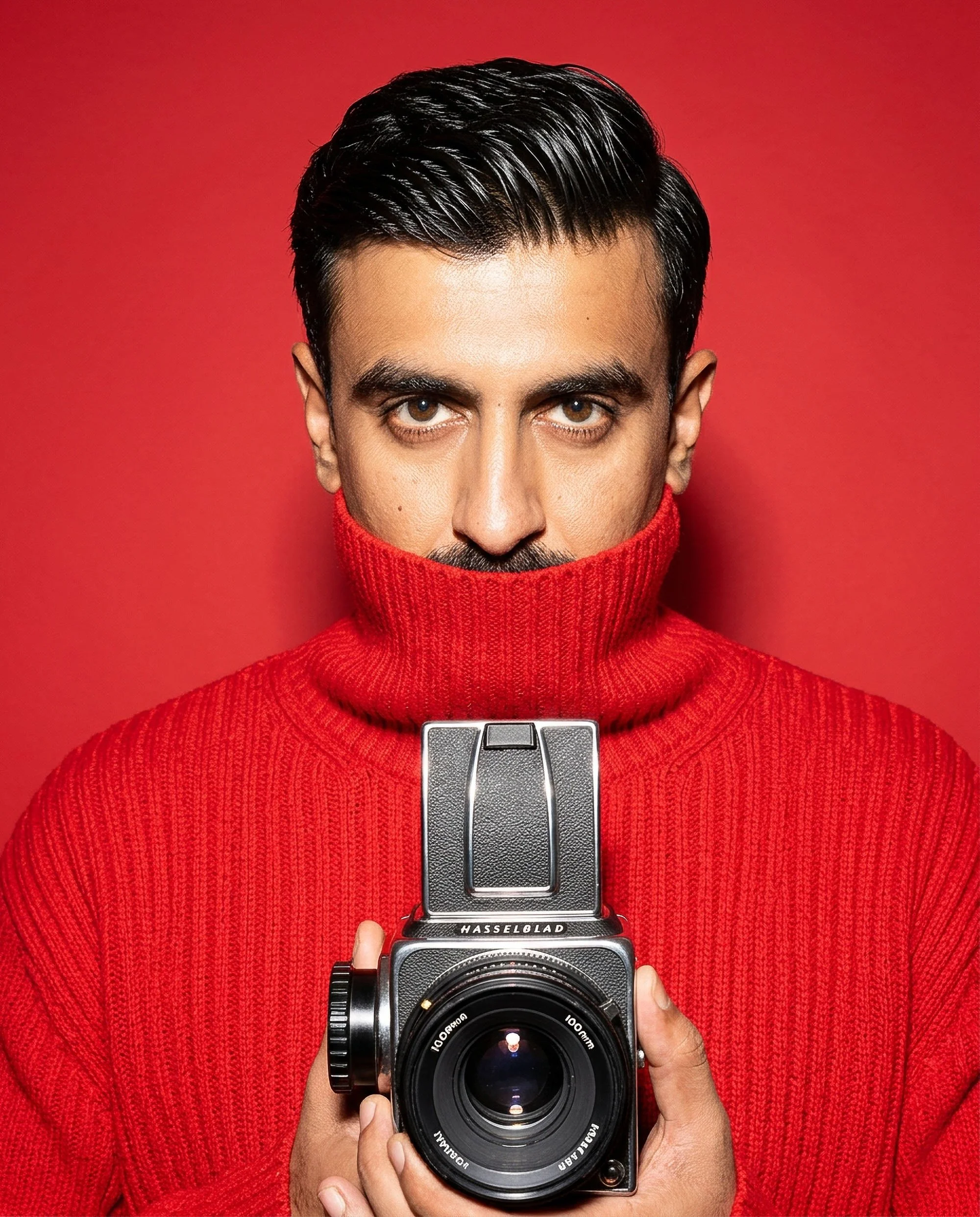 A person wearing a red turtleneck sweater holding a Hasselblad camera in front of a matching red background.