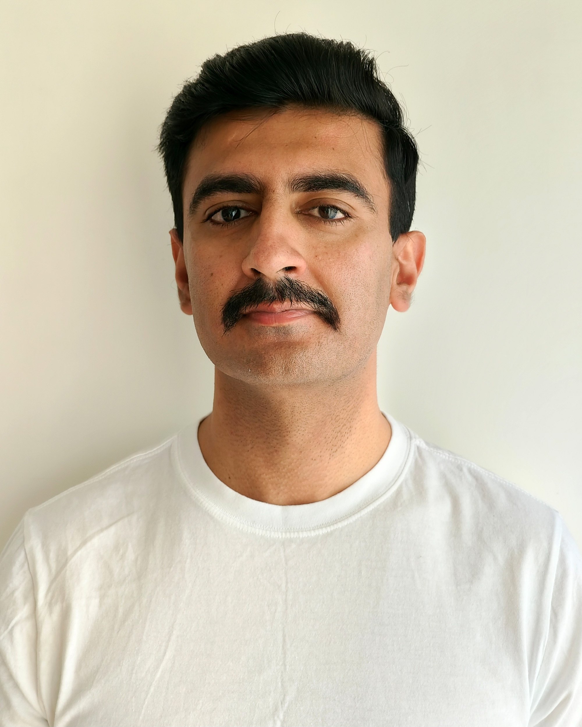 Close-up of a man with dark hair, mustache, and light skin, wearing a white shirt, standing against a plain light-colored wall.