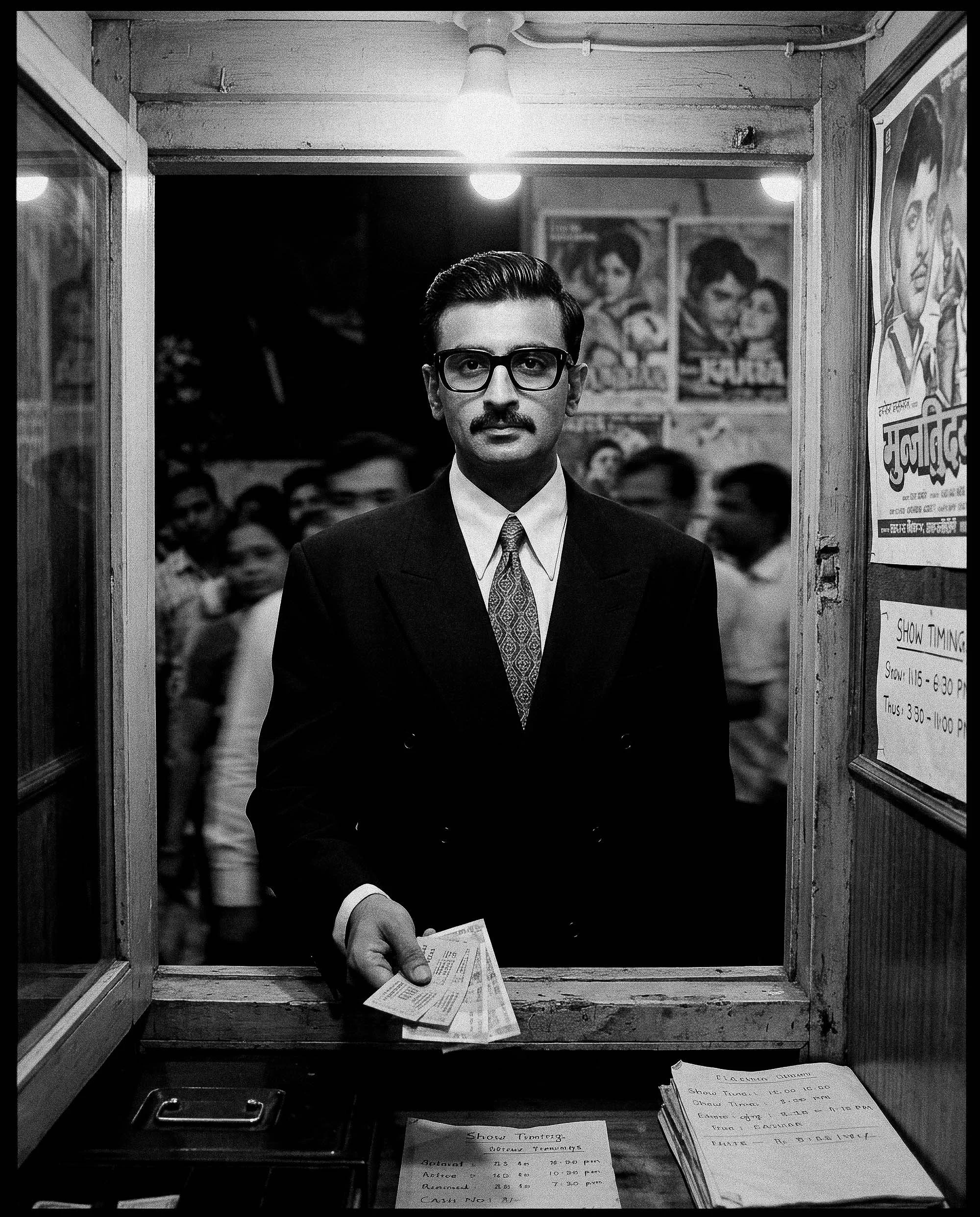 Black and white photo of a man with glasses, a mustache, wearing a suit and tie, standing behind a counter with his hand extended holding a ticket or receipt. There are posters of people on the wall behind him, and papers on the counter with show tim